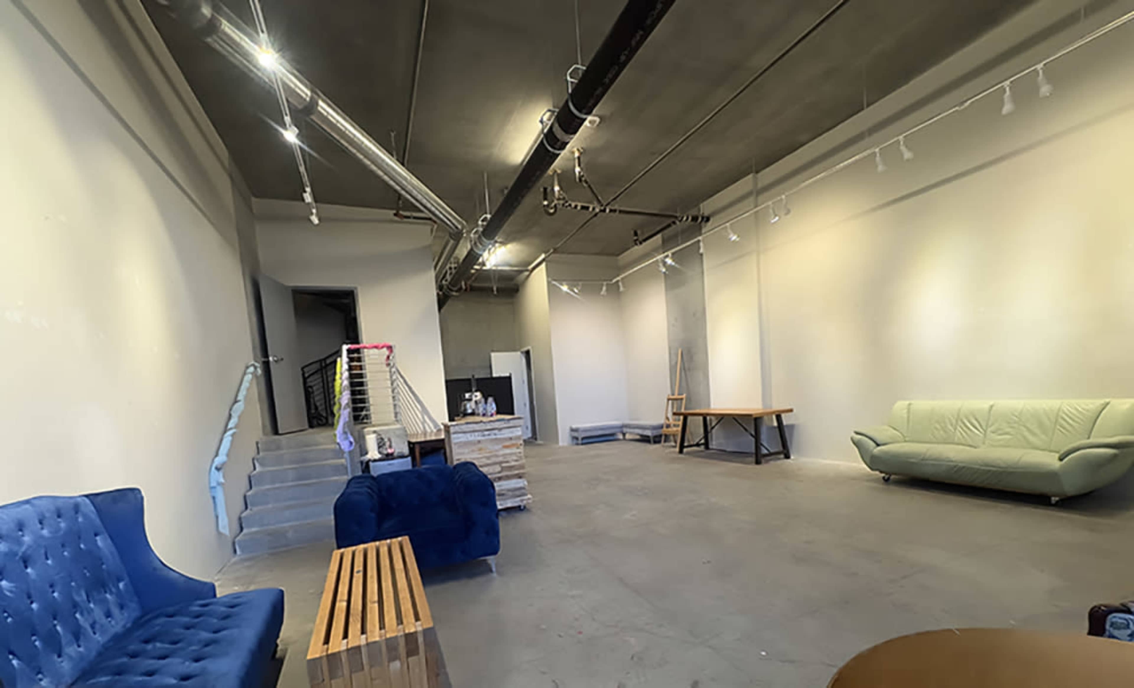 The image shows a spacious, empty room with concrete flooring, two sofas, a wooden table, and a staircase leading to another level.