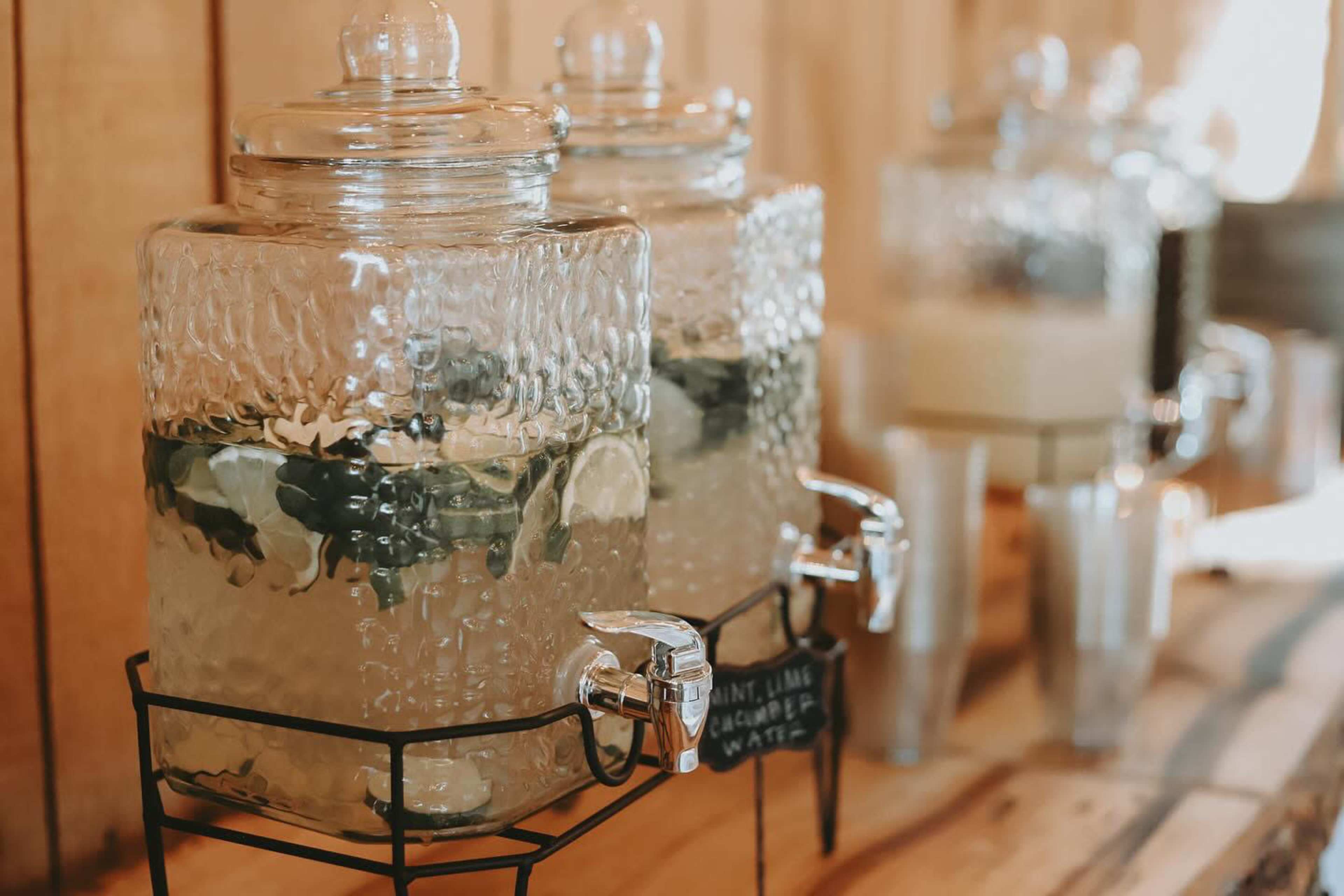Two glass beverage dispensers filled with infused water, featuring mint, lime, and cucumber, are placed on a wooden table.