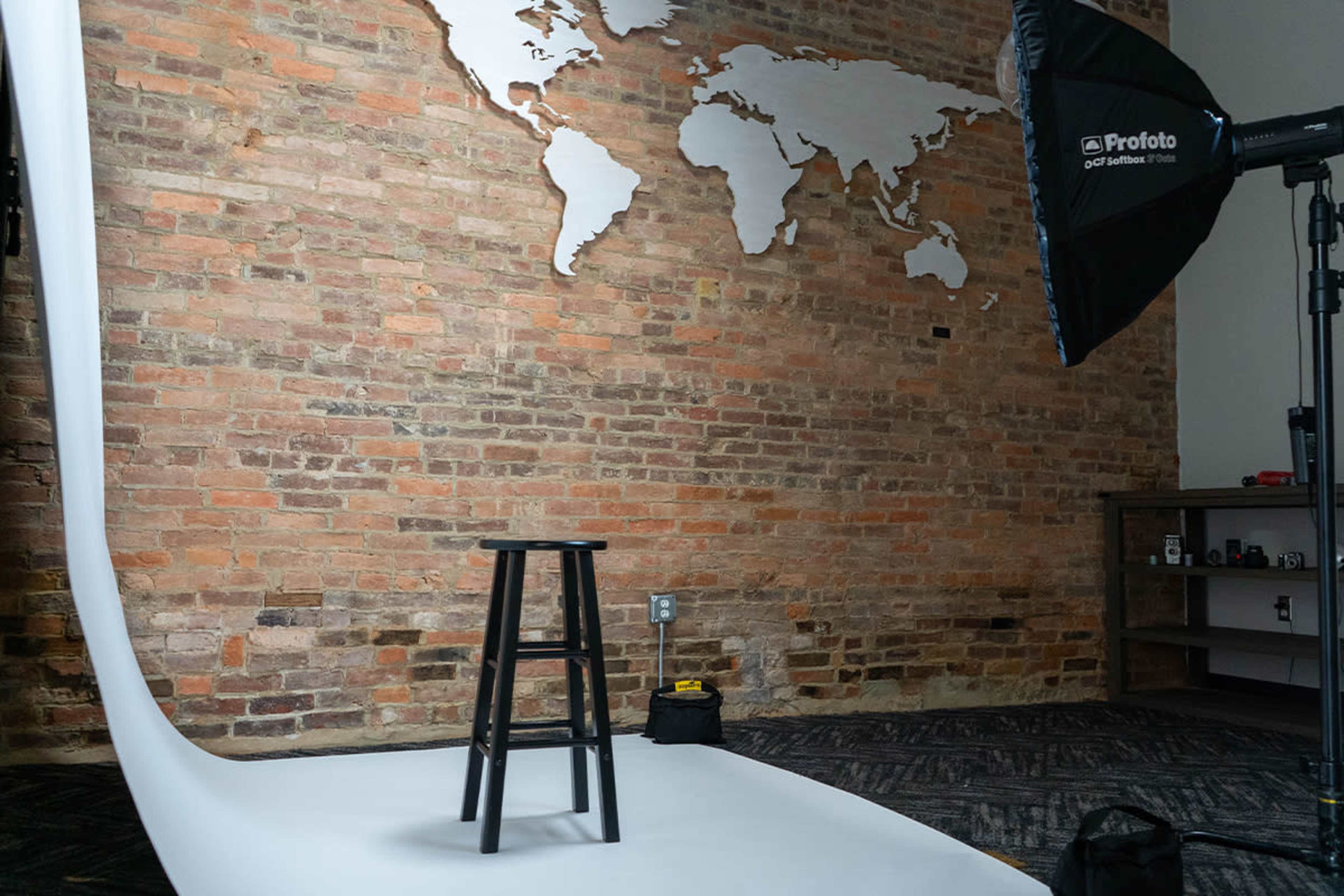The image shows a photography studio with a wooden stool on a white backdrop, against a brick wall featuring a world map silhouette.