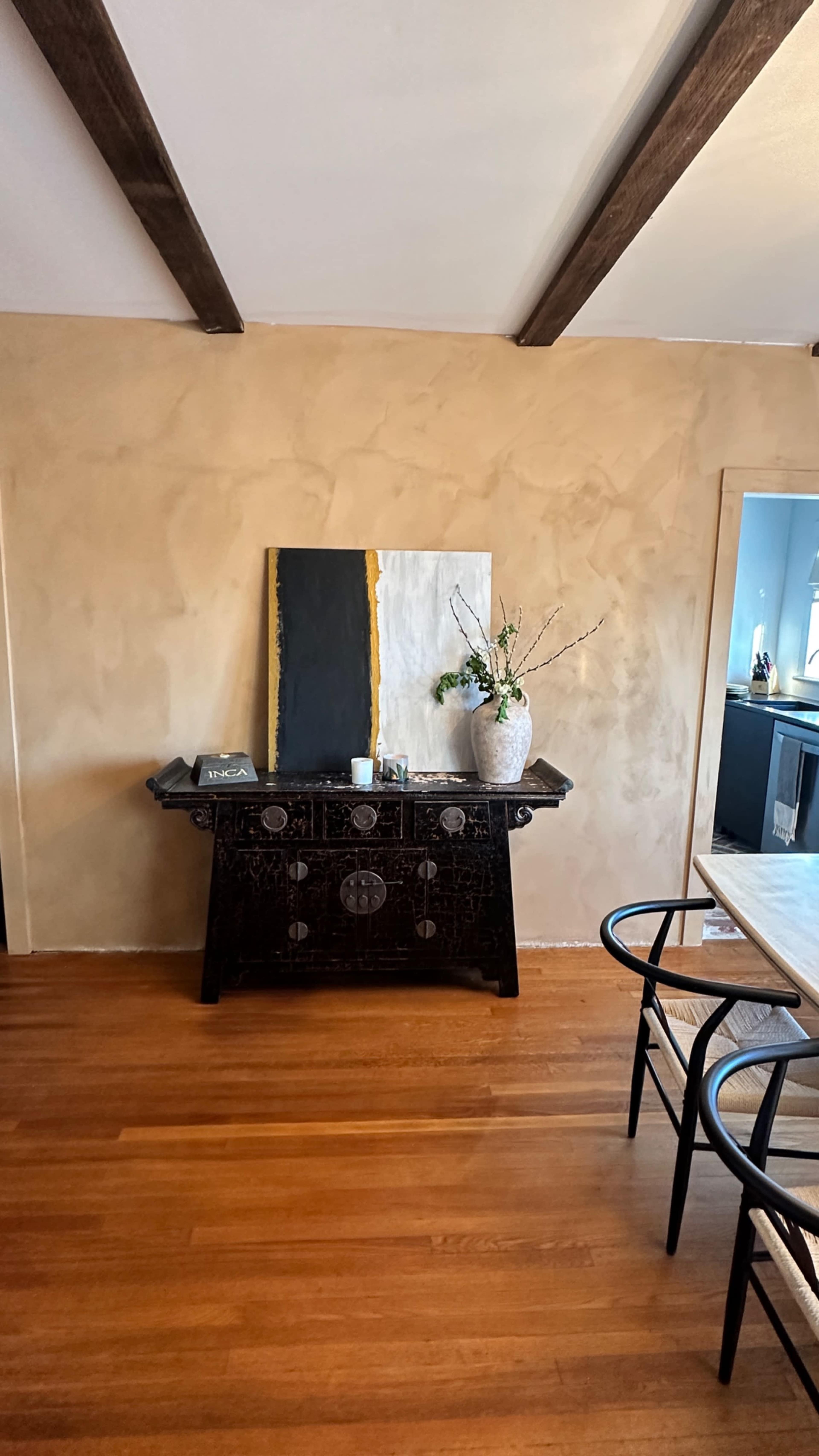 The image showcases a room with a wooden floor, a black chest of drawers against a textured beige wall, and a large abstract painting above it, complemented by a vase of branches.