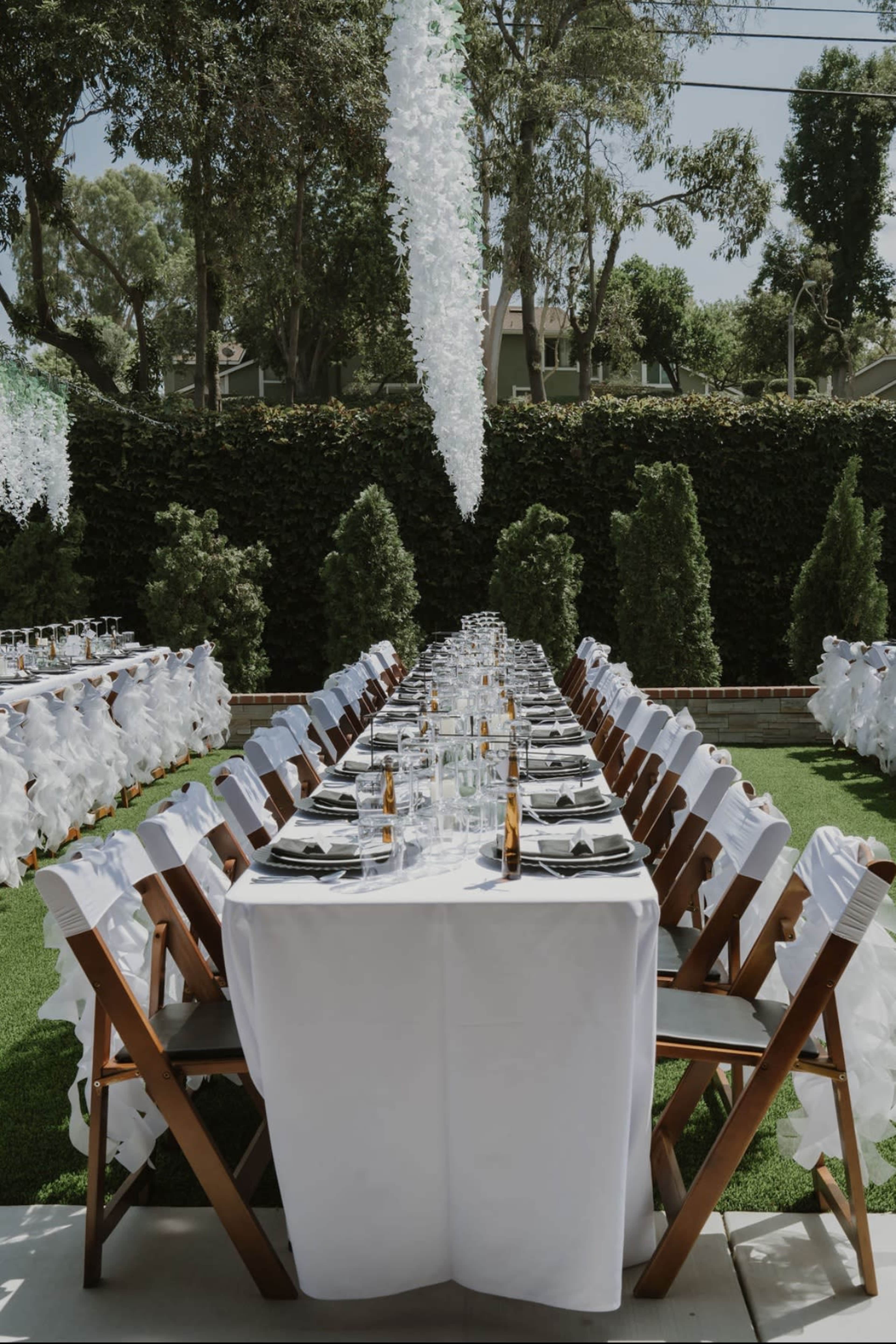 A long outdoor dining table is set with white tablecloths and black napkins, surrounded by neatly arranged chairs, with greenery in the background.