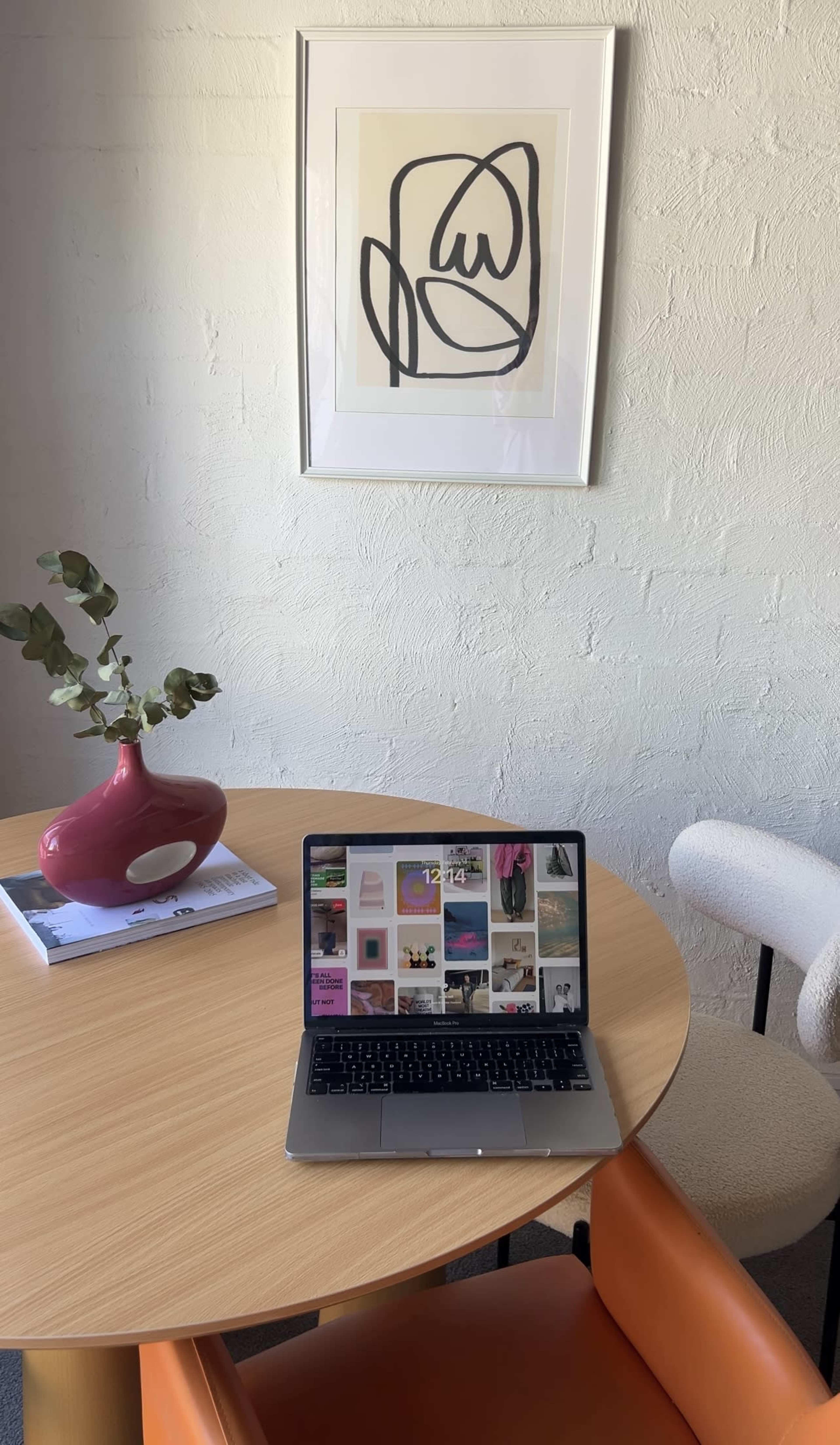 A laptop displaying a grid of images is placed on a wooden table next to a pink vase with a single stem plant, with a piece of abstract art hanging on the wall above.