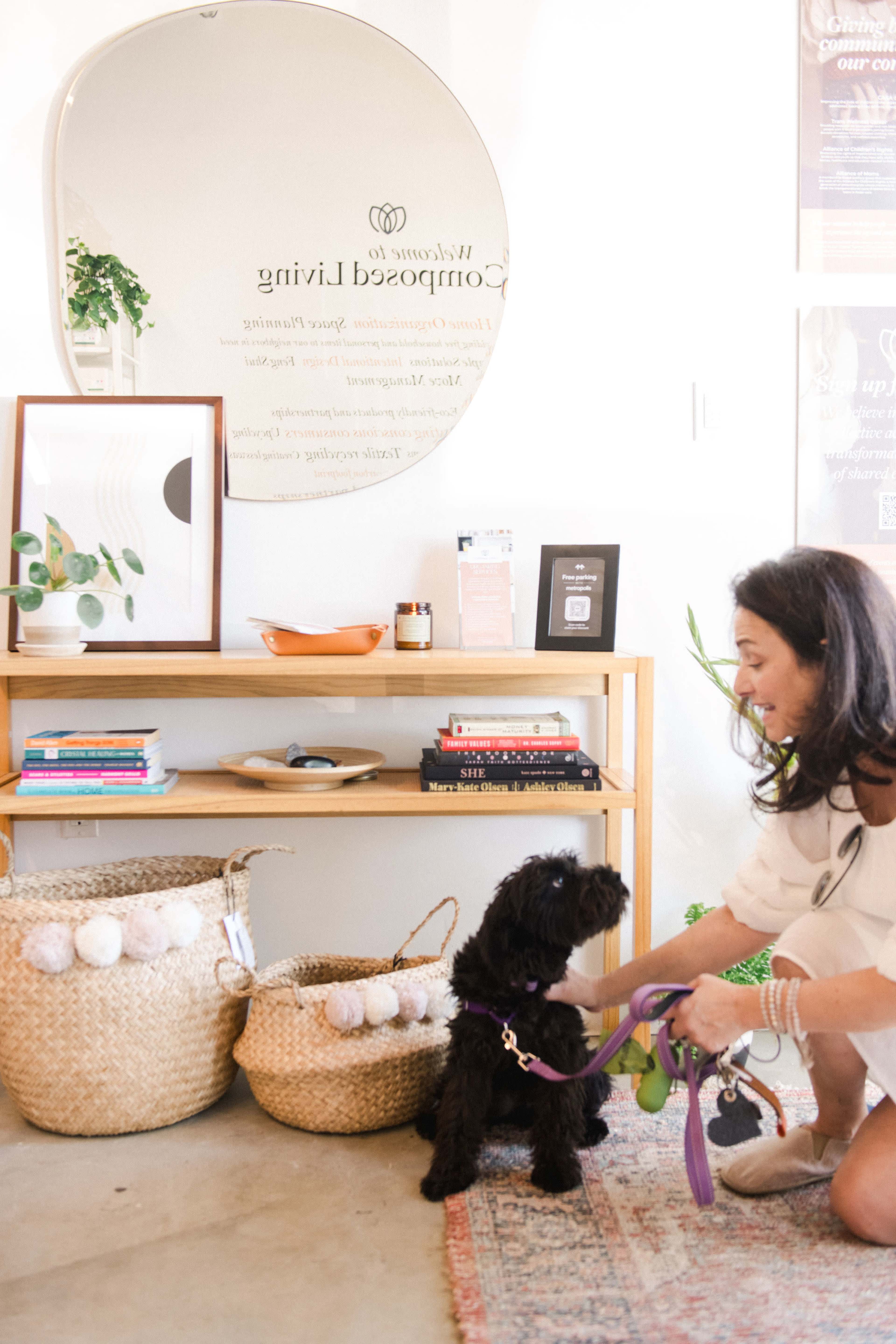 A woman kneels in front of a black dog, preparing to attach a leash, while standing next to a wooden shelf filled with books and decorative items.
