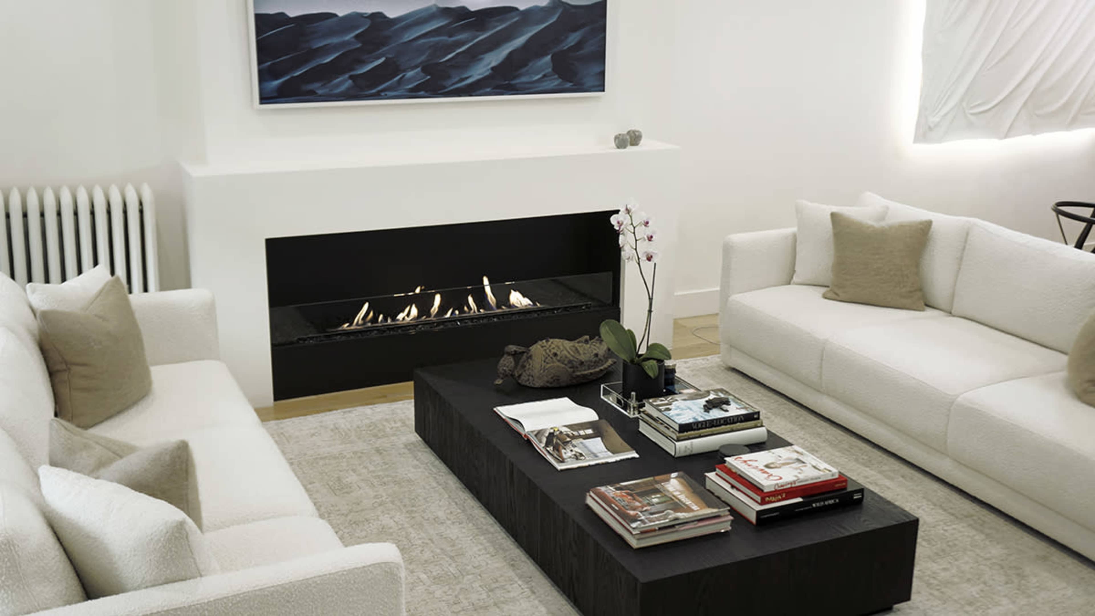 A modern living room features two white sofas facing a black coffee table, with a fireplace installed in a sleek wall and artworks hanging above.