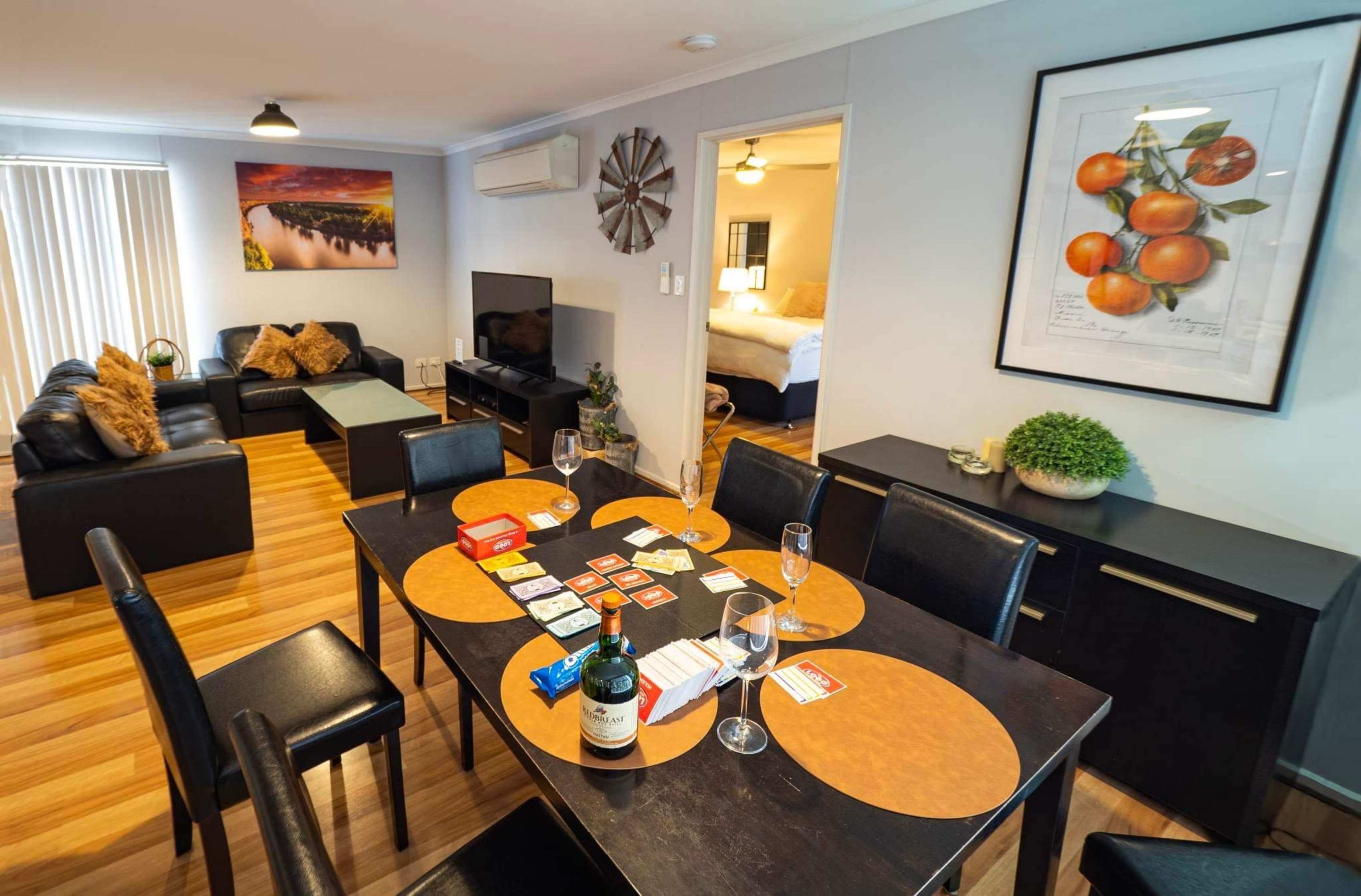 The image shows a cozy living area with a dining table set for a meal, featuring a bottle of wine and board games, alongside a lounge area with black sofas and a television.