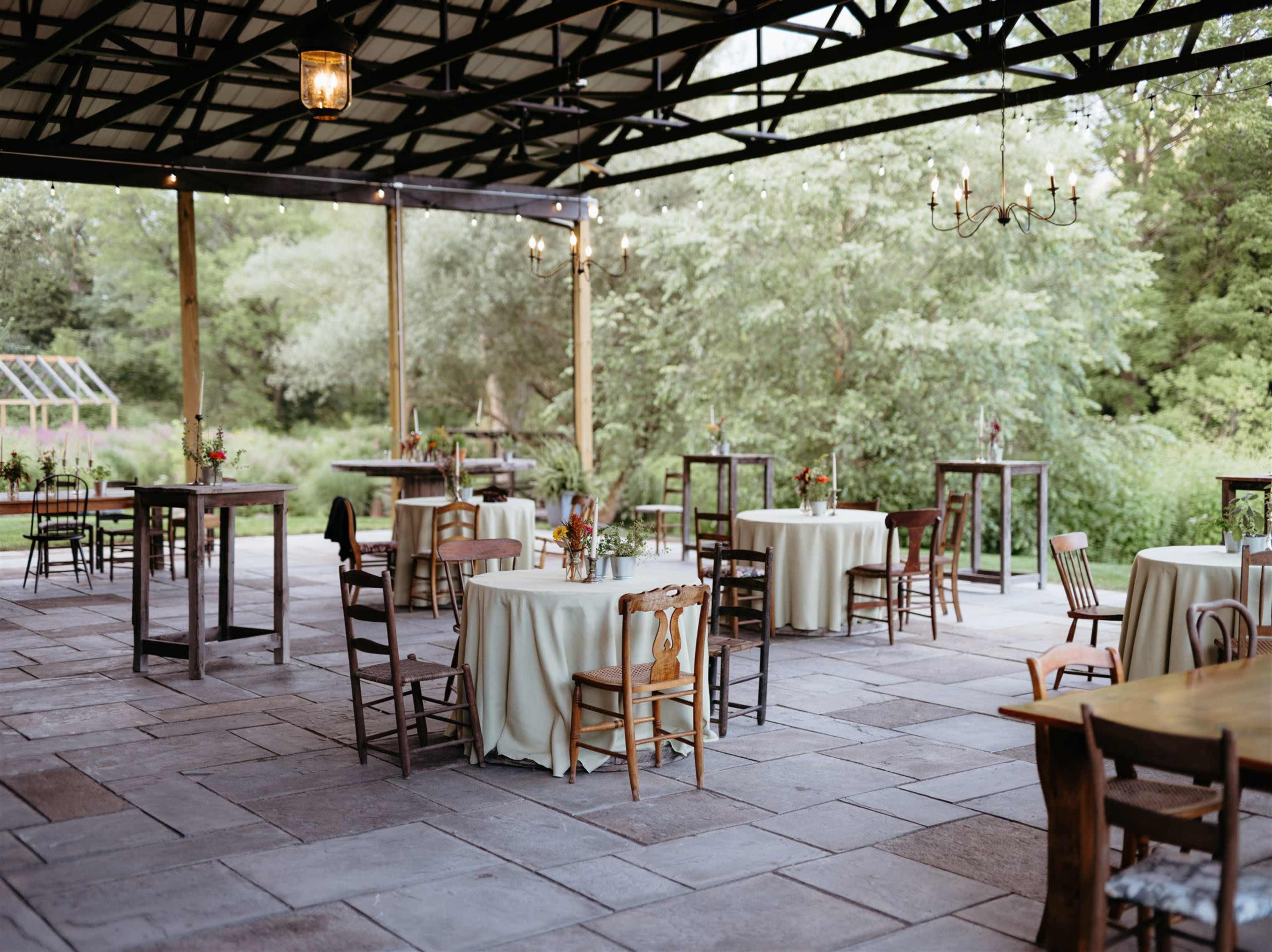 A spacious outdoor pavilion features several tables with light-colored tablecloths arranged on a stone floor, surrounded by greenery.
