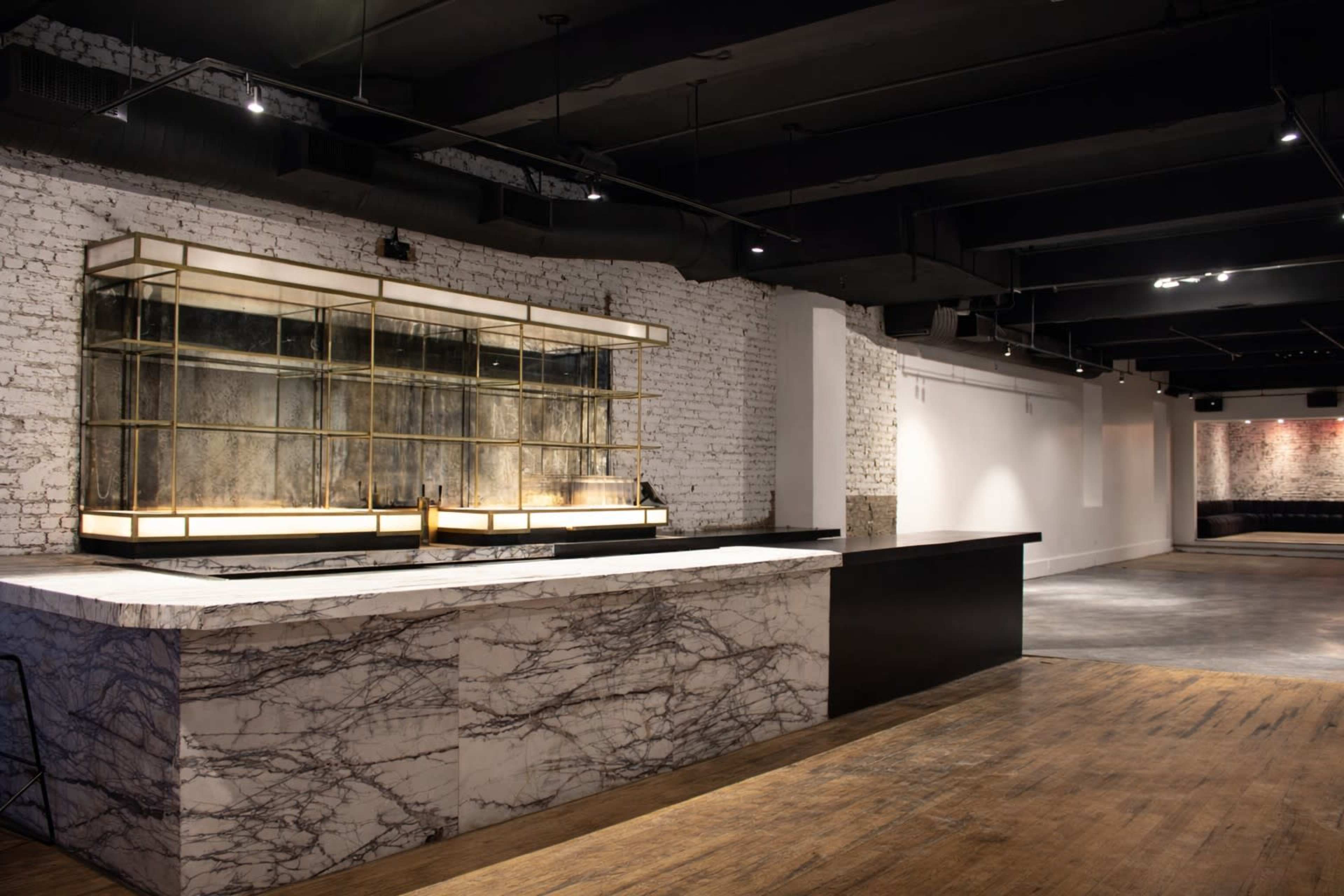 The image shows a modern, minimalist interior featuring a marble bar counter with a glass display case against a backdrop of exposed white brick walls and wooden flooring.