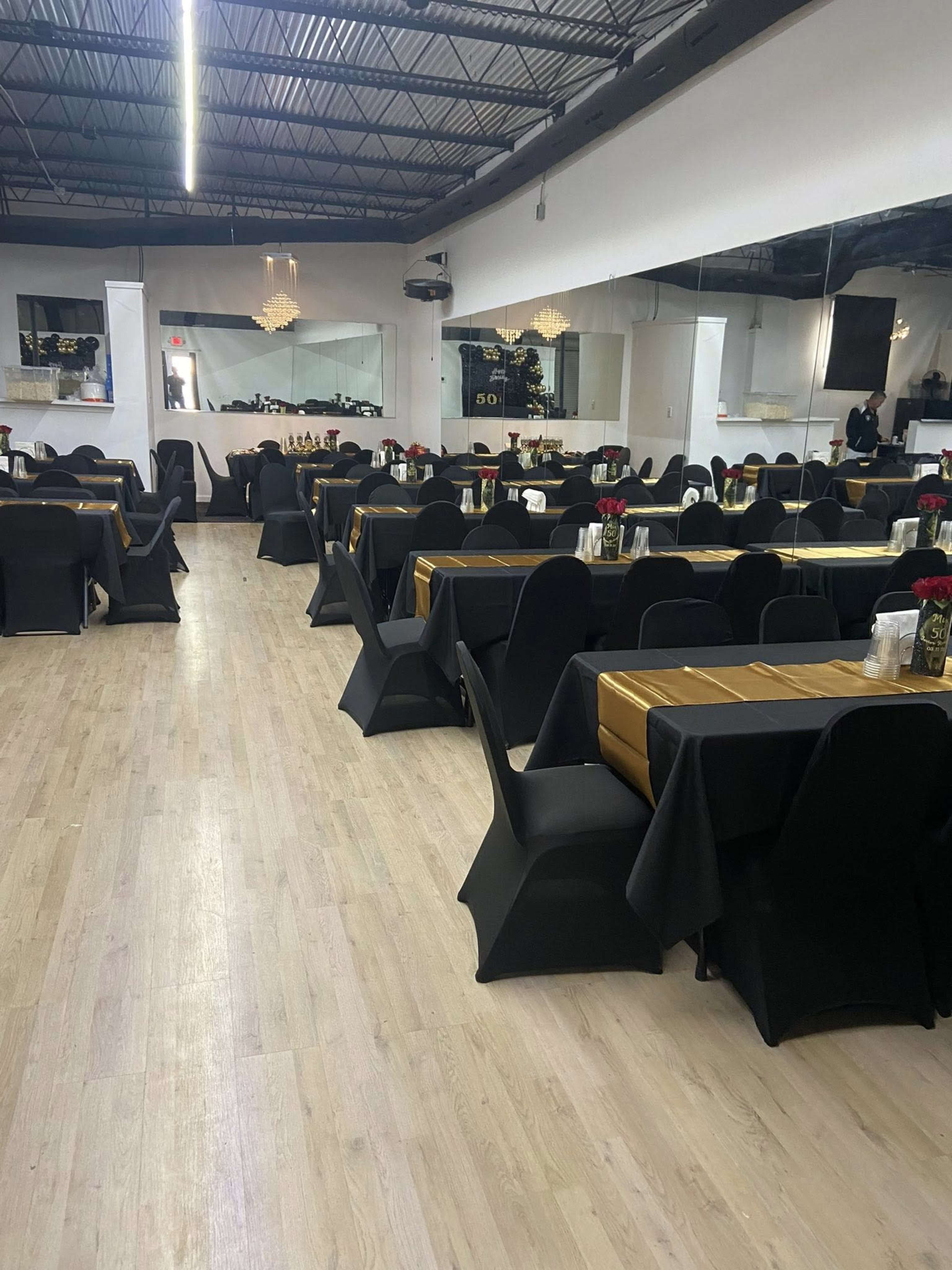 A spacious event hall is set up with tables covered in black and gold tablecloths, featuring centerpieces with flowers and candles.