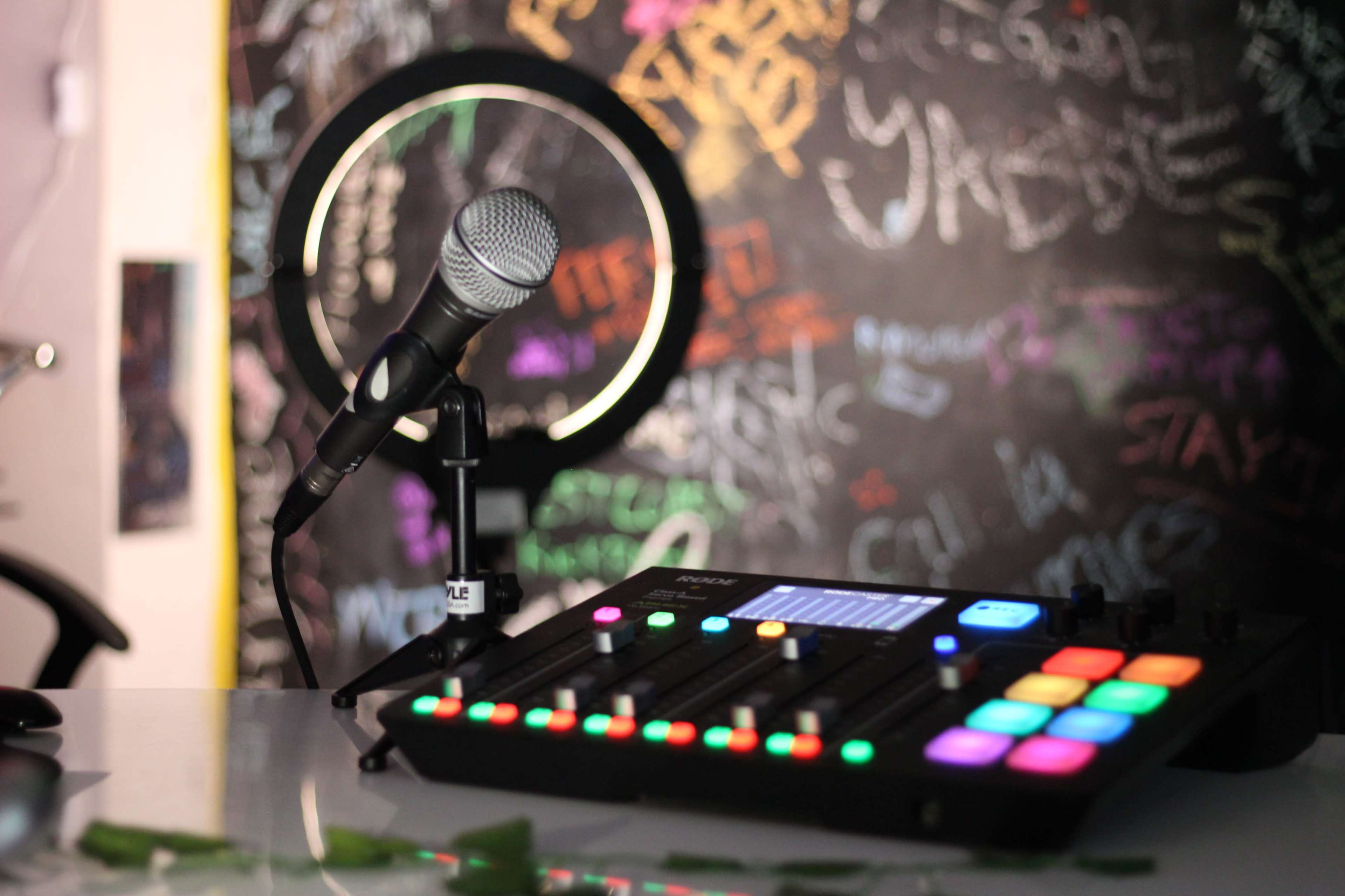 A microphone stands on a stand next to a colorful audio mixer, set against a wall covered in chalk graffiti.