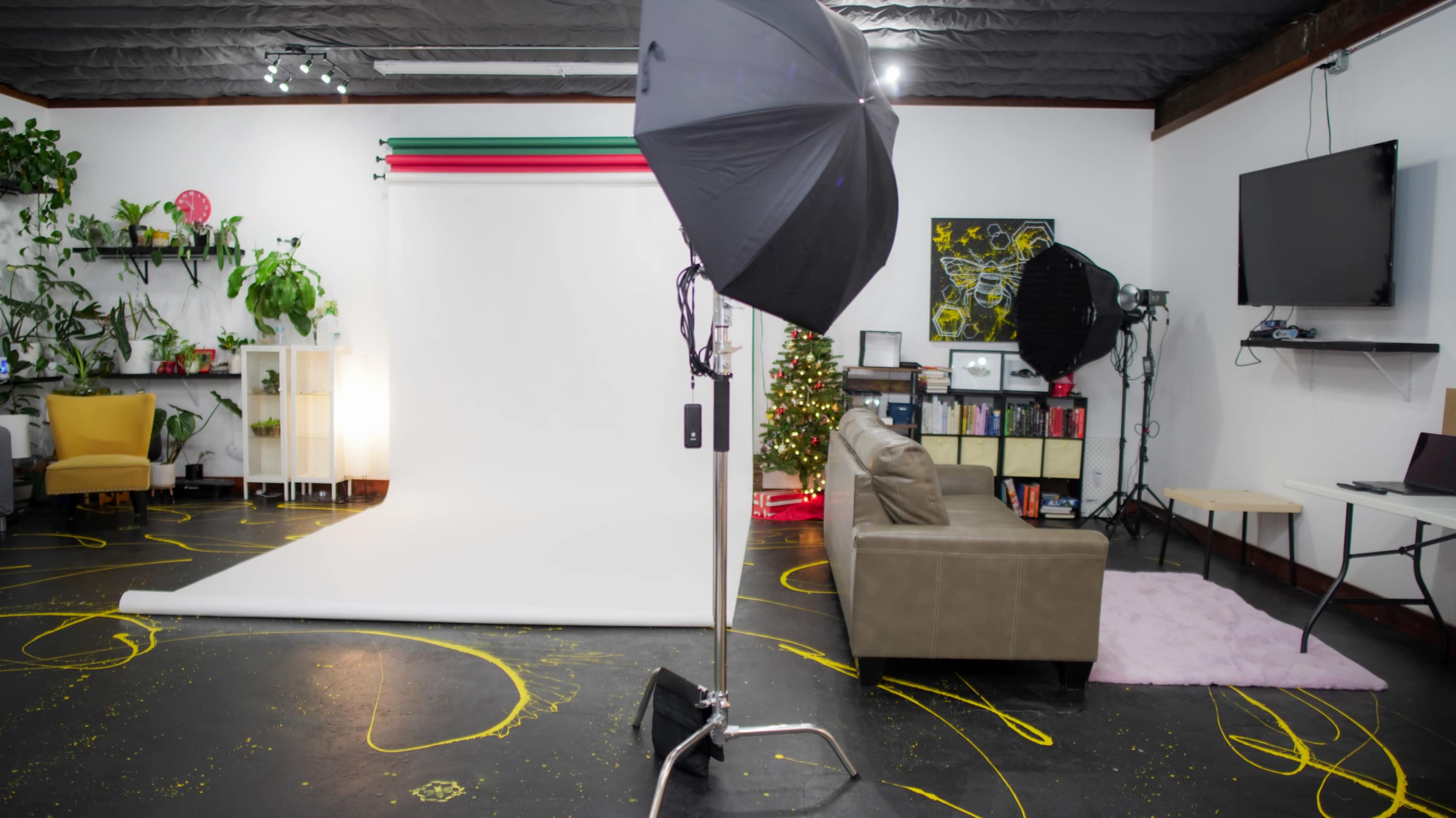 The image depicts a photography studio featuring a white backdrop, an umbrella light, a beige sofa, and a decorated Christmas tree in the background.