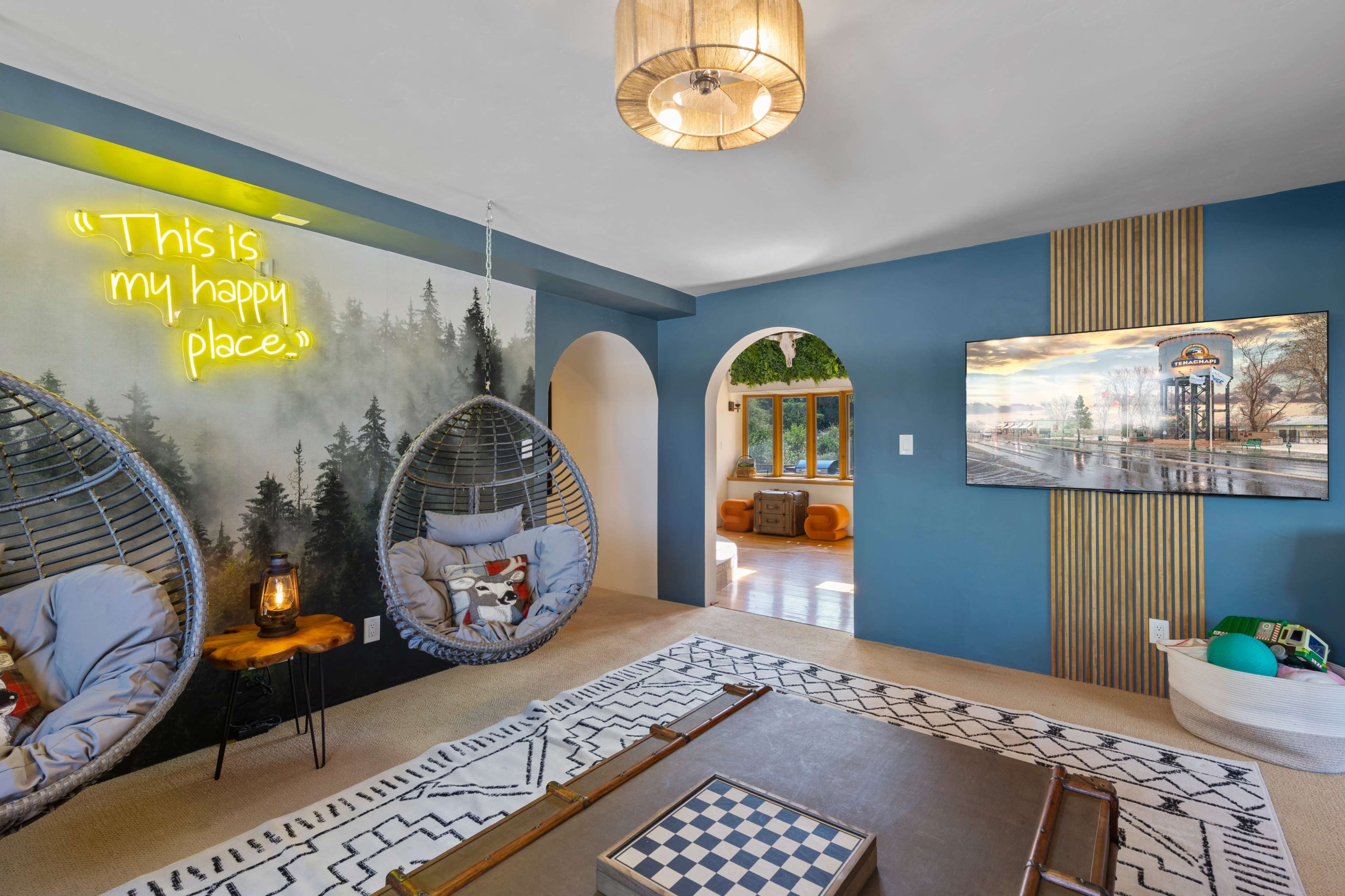 A cozy living room featuring two hanging chairs, a neon sign, a large television, and a patterned rug.