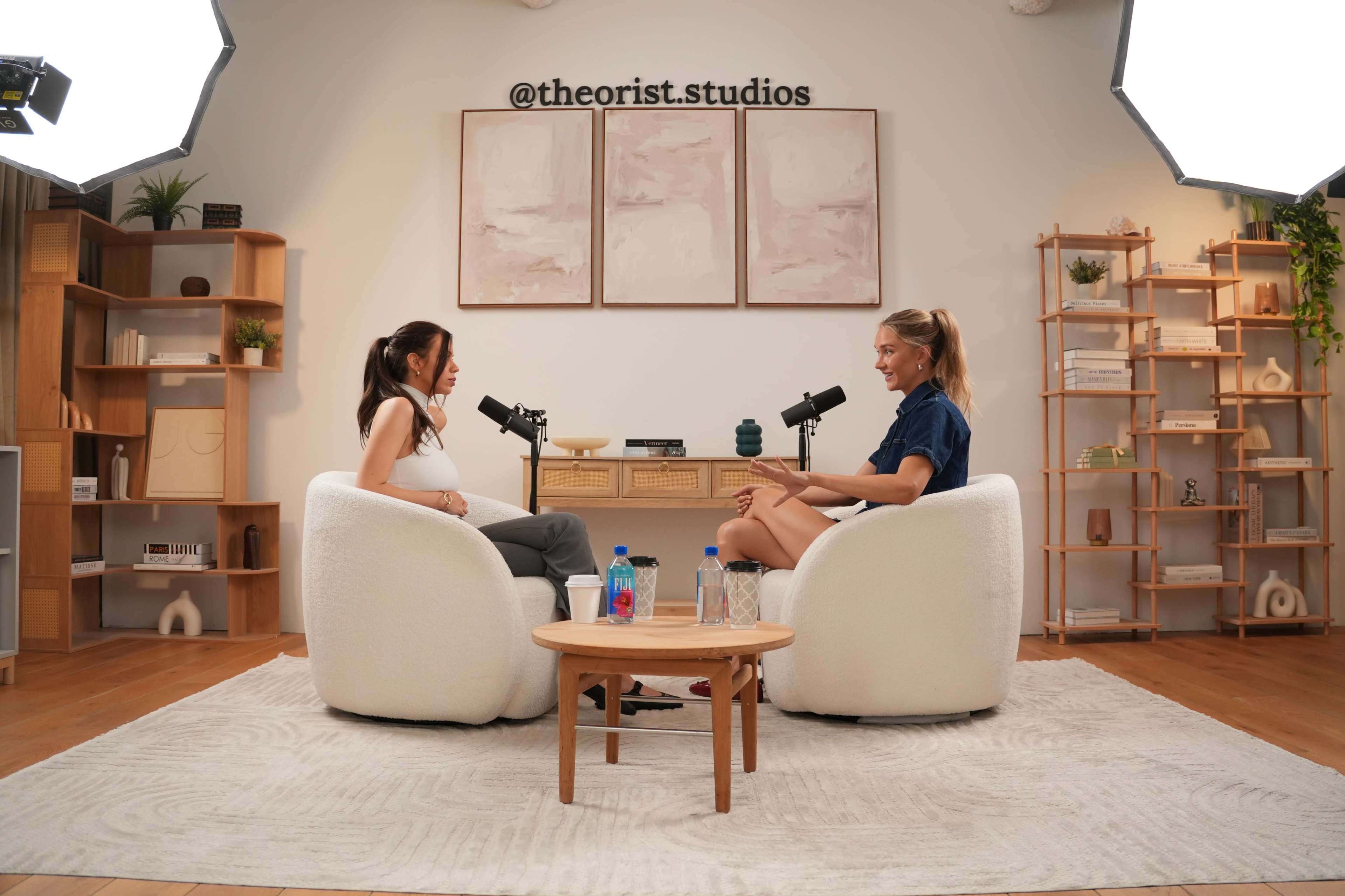 Two women are seated in comfortable chairs facing each other in a well-lit studio, equipped with microphones, decorative shelves, and a neutral-toned rug.
