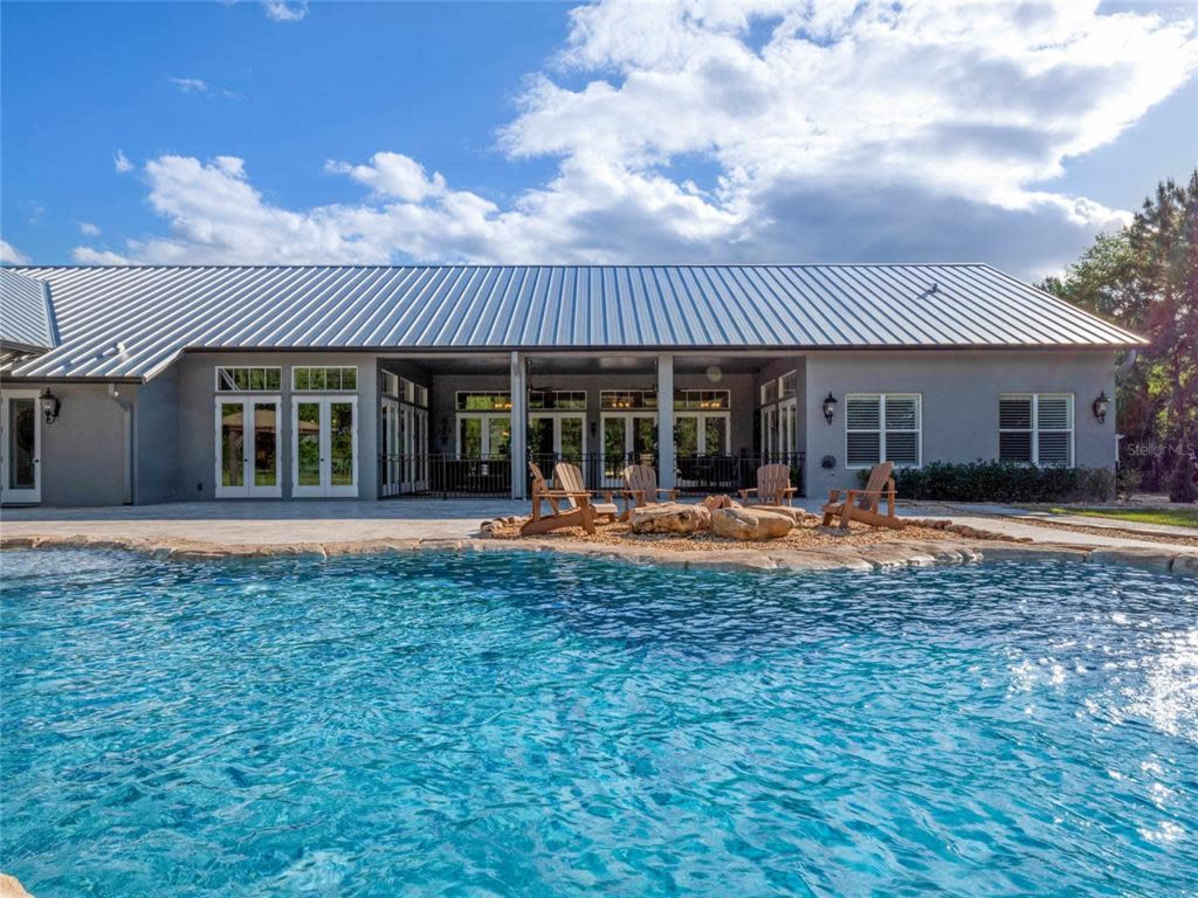 A modern house with a metal roof overlooks a swimming pool with stone landscaping and wooden lounge chairs.