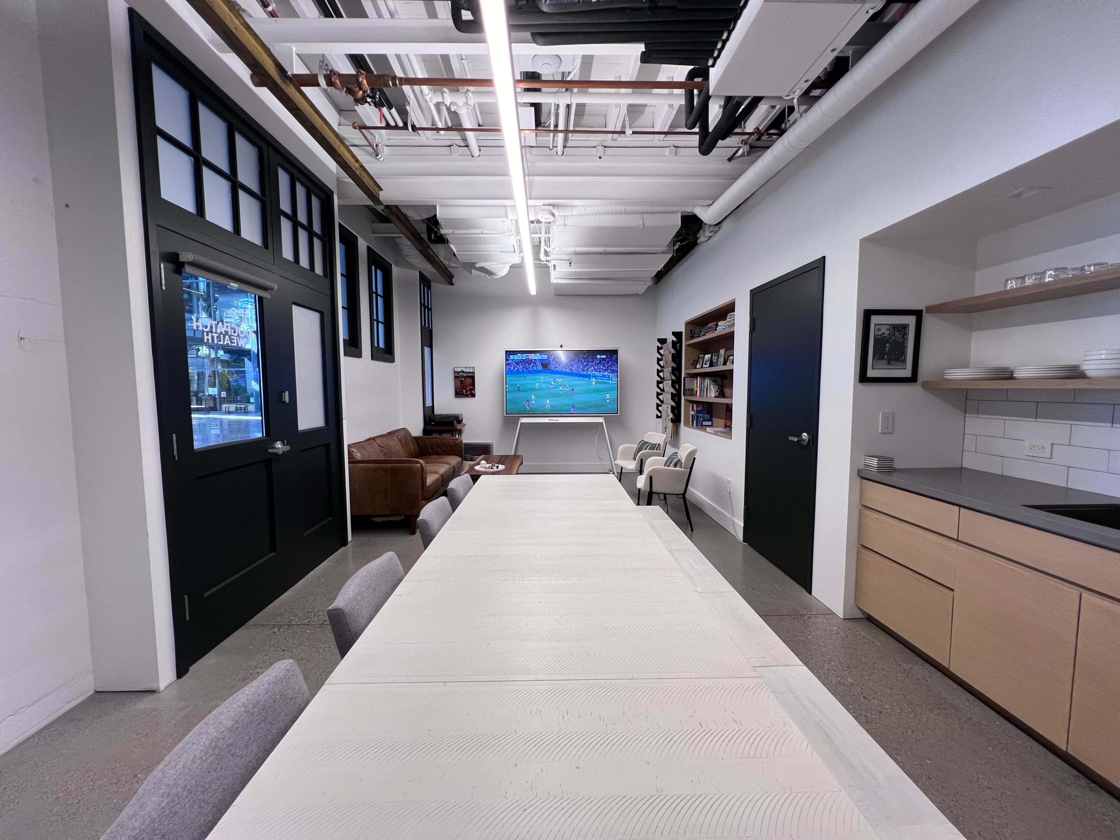 A spacious room features a long dining table, a couch, and a wall-mounted TV displaying a sports game.
