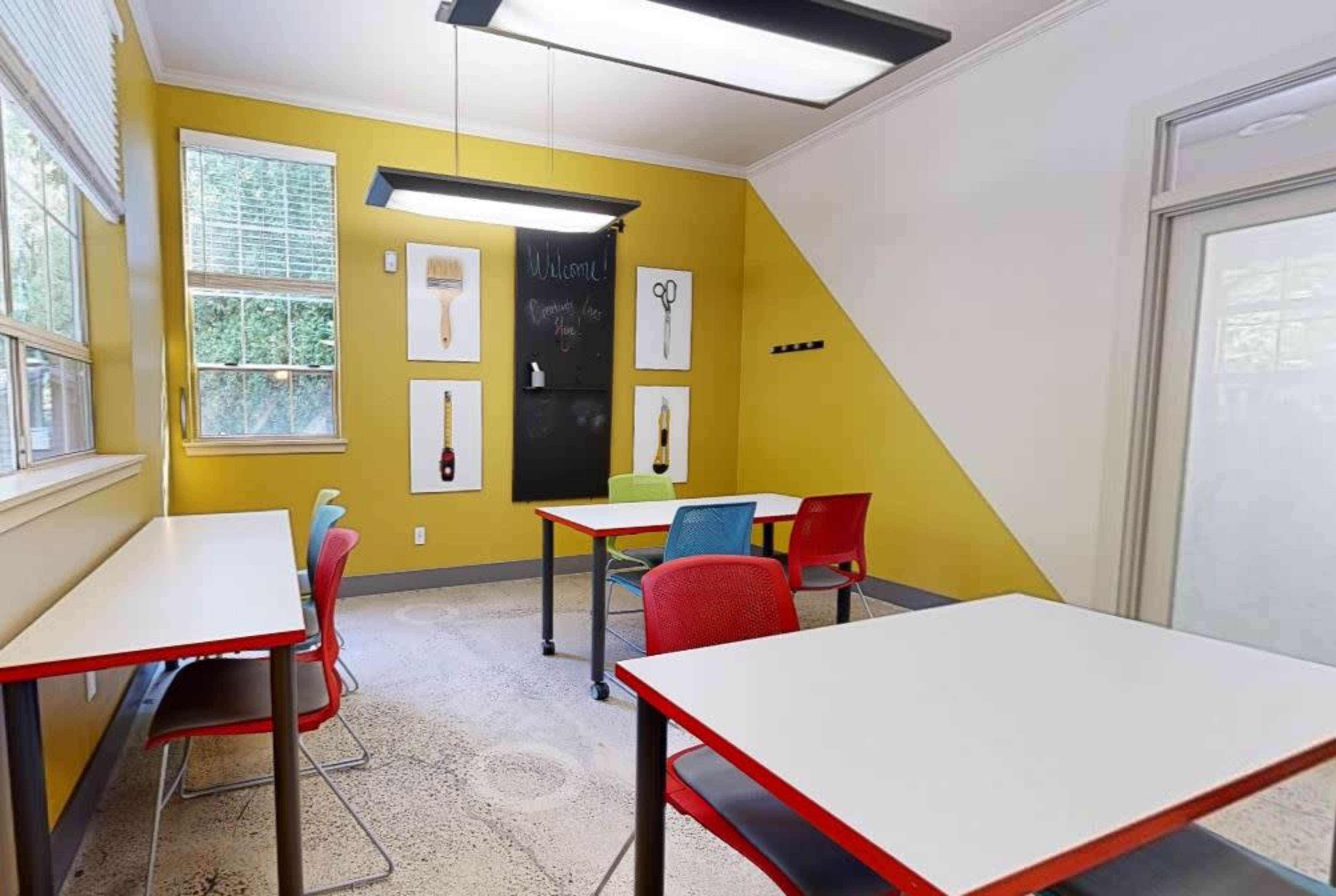 A classroom features bright yellow walls, two tables with red and gray chairs, and artwork hanging on the walls.