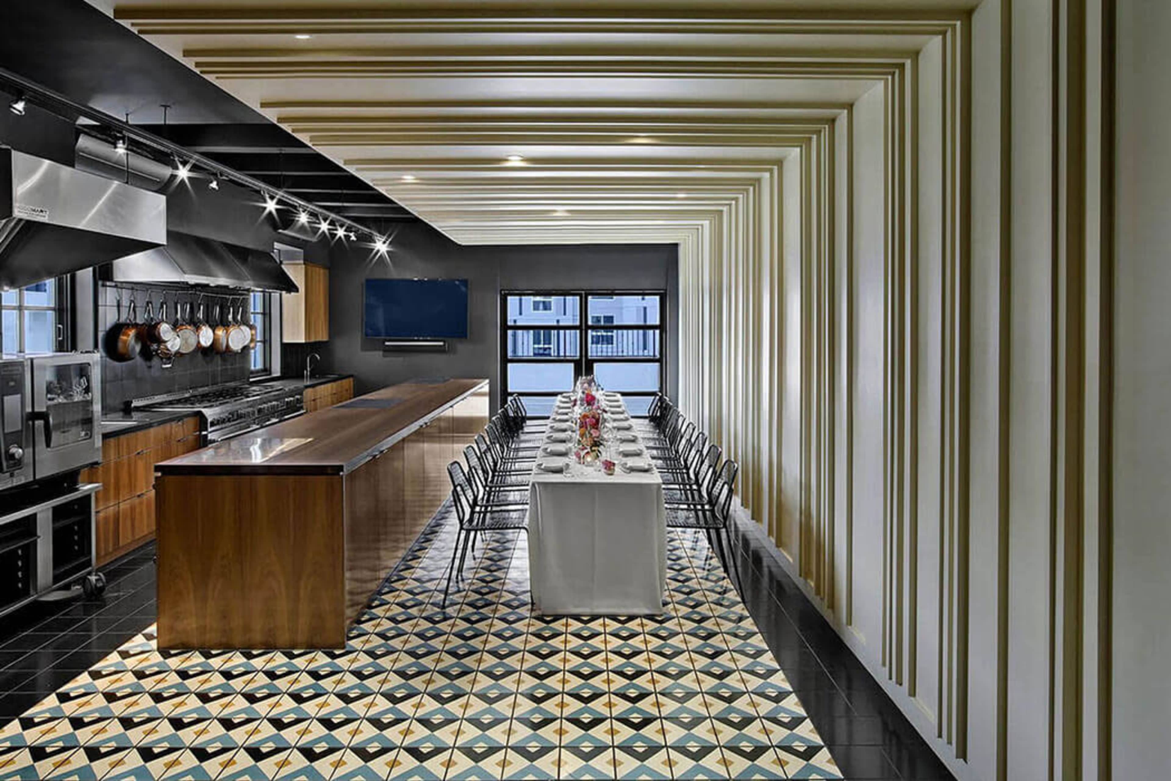The image shows a modern kitchen and dining area featuring a long table set for a meal, surrounded by sleek chairs, with a geometric patterned floor and a backdrop of wooden cabinetry and stainless steel appliances.