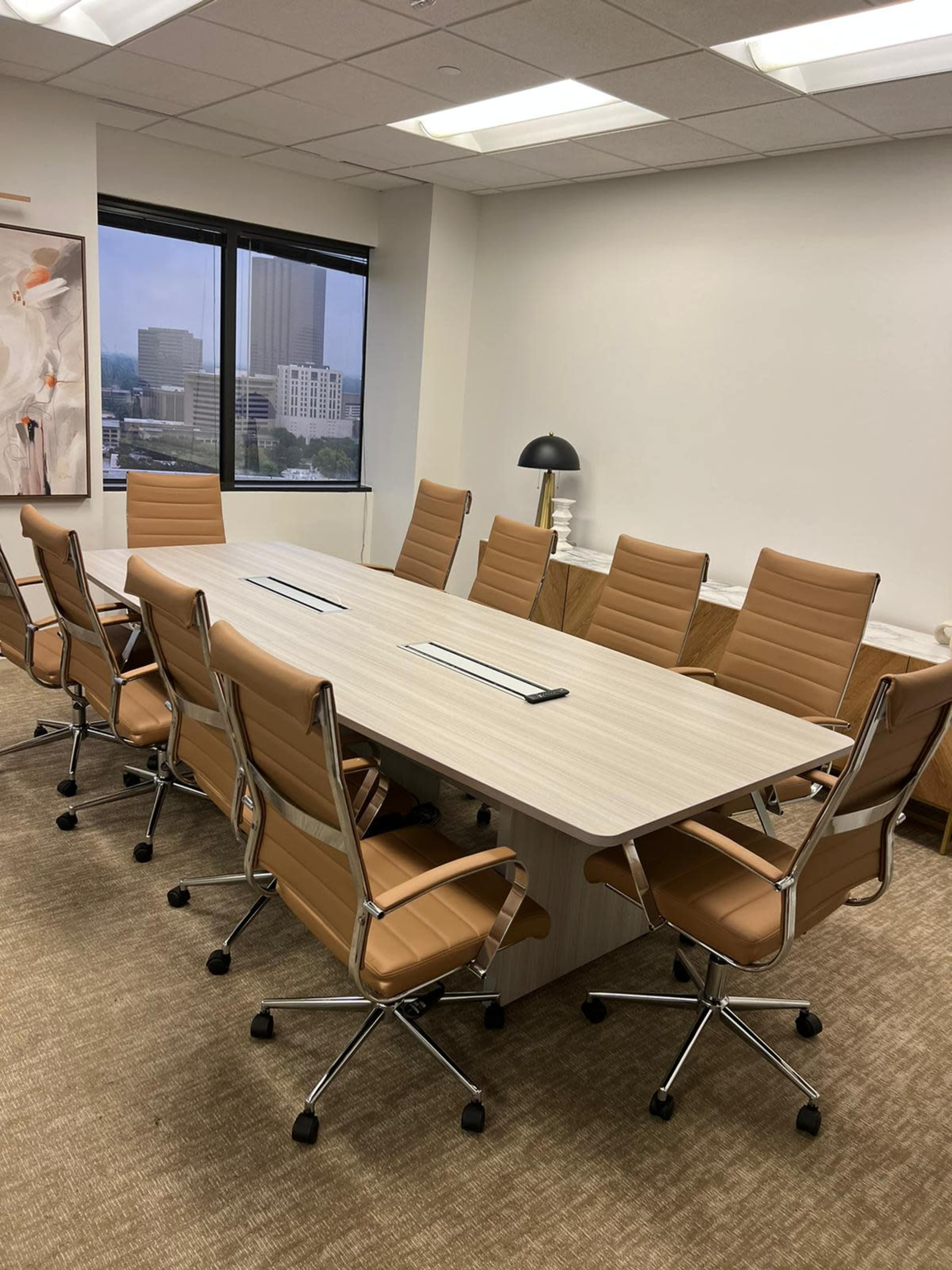A modern conference room with a long wooden table surrounded by eight beige rolling chairs, and large windows providing a view of a city skyline.