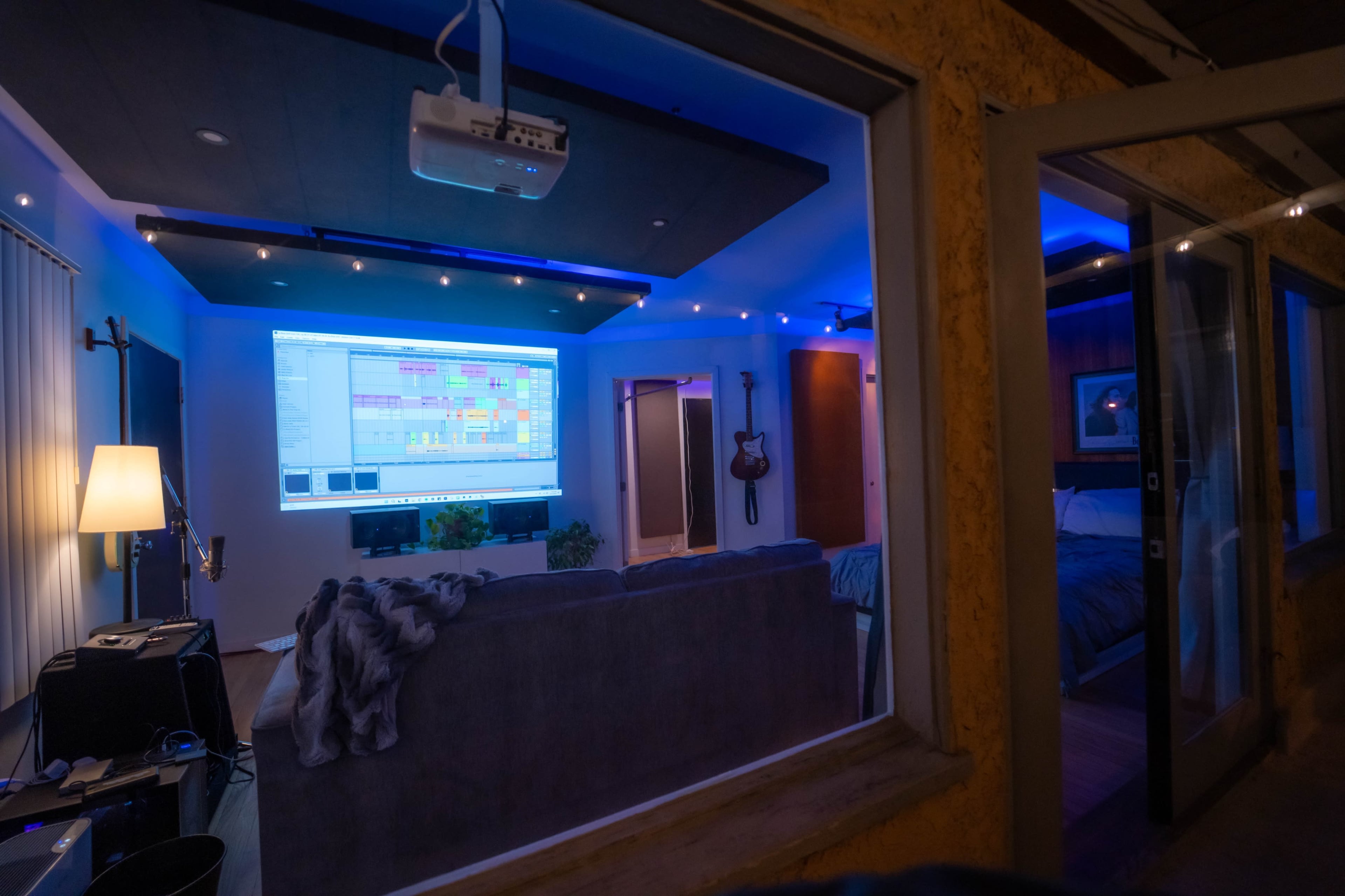 A cozy living room with a large projection screen displaying music software, illuminated by blue ambient lighting and a guitar hanging on the wall.