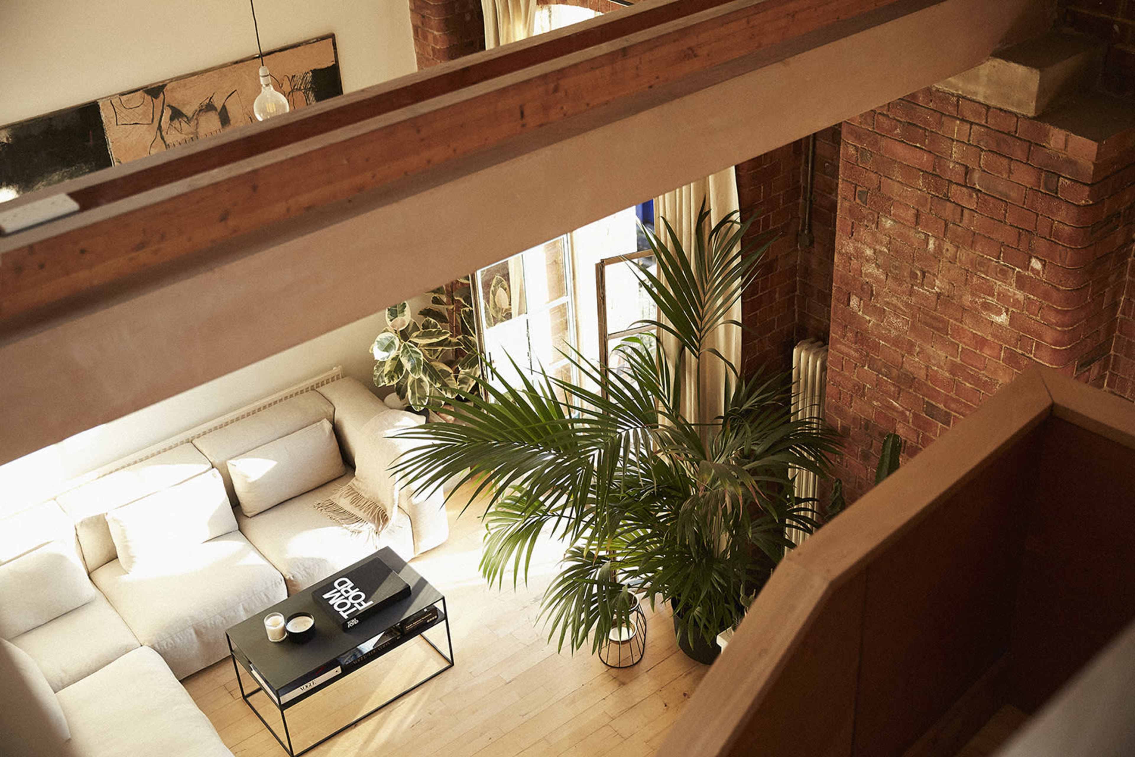 A spacious living room features a beige sectional sofa, a coffee table with decorative items, and a large potted plant, all set against exposed brick walls and large windows.