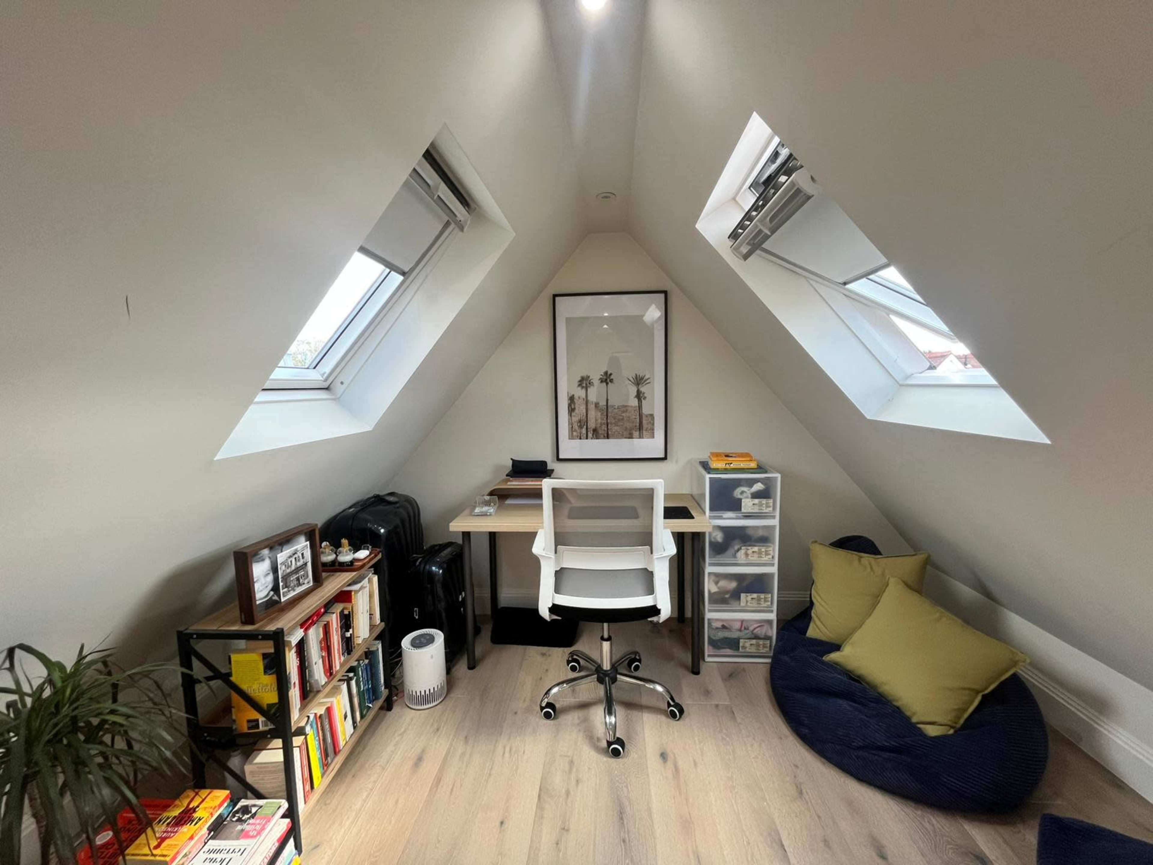 The image depicts a small attic space with a desk and chair positioned under two slanted windows, surrounded by shelves of books, a cozy beanbag, and a plant in one corner.