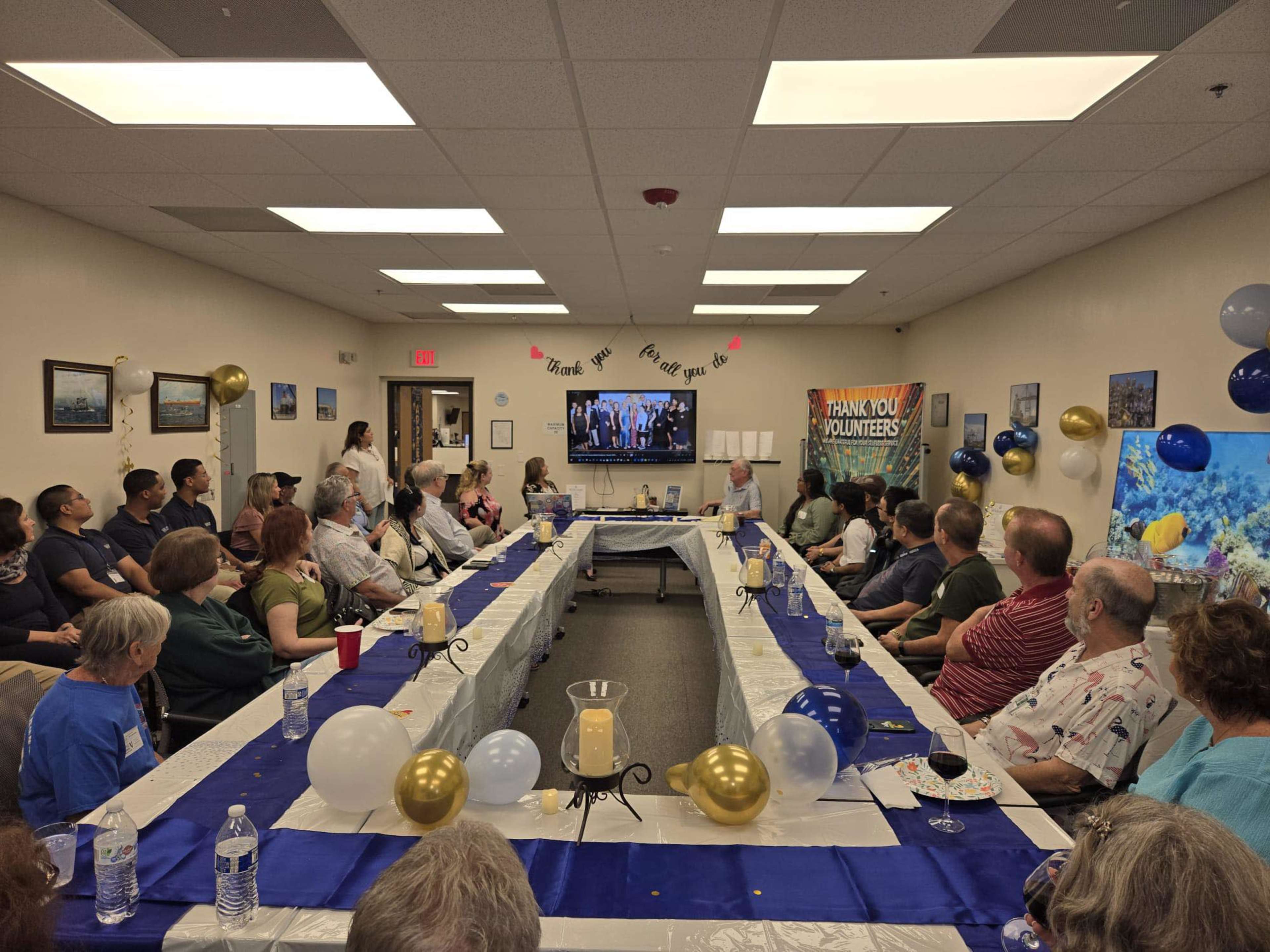 A group of people gathers around a long table decorated with balloons and a banner to celebrate and thank volunteers, while a presentation plays on a screen.