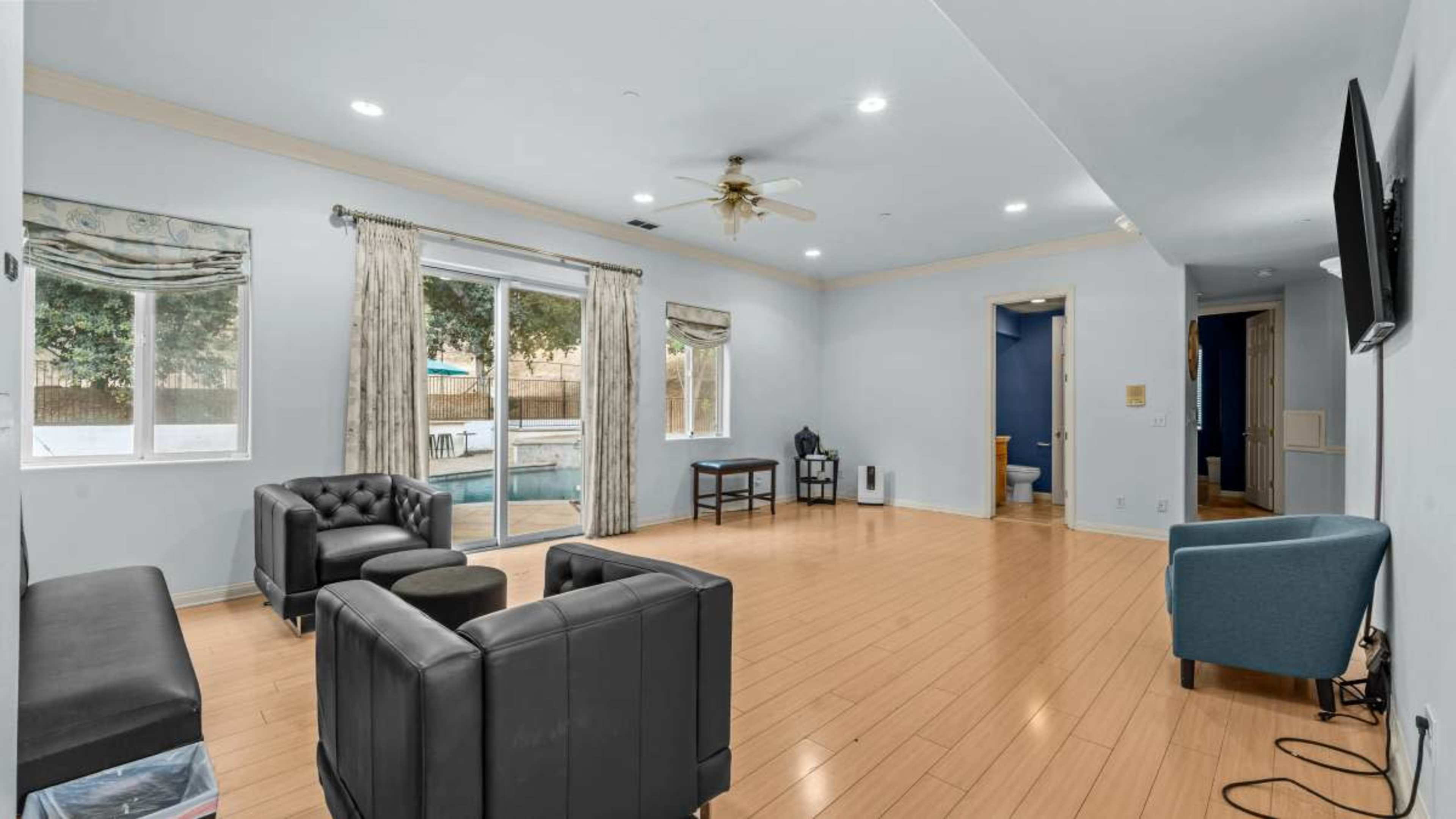 The image shows a spacious living room with bamboo flooring, two black armchairs, a blue chair, large windows, and a doorway leading to a bathroom.