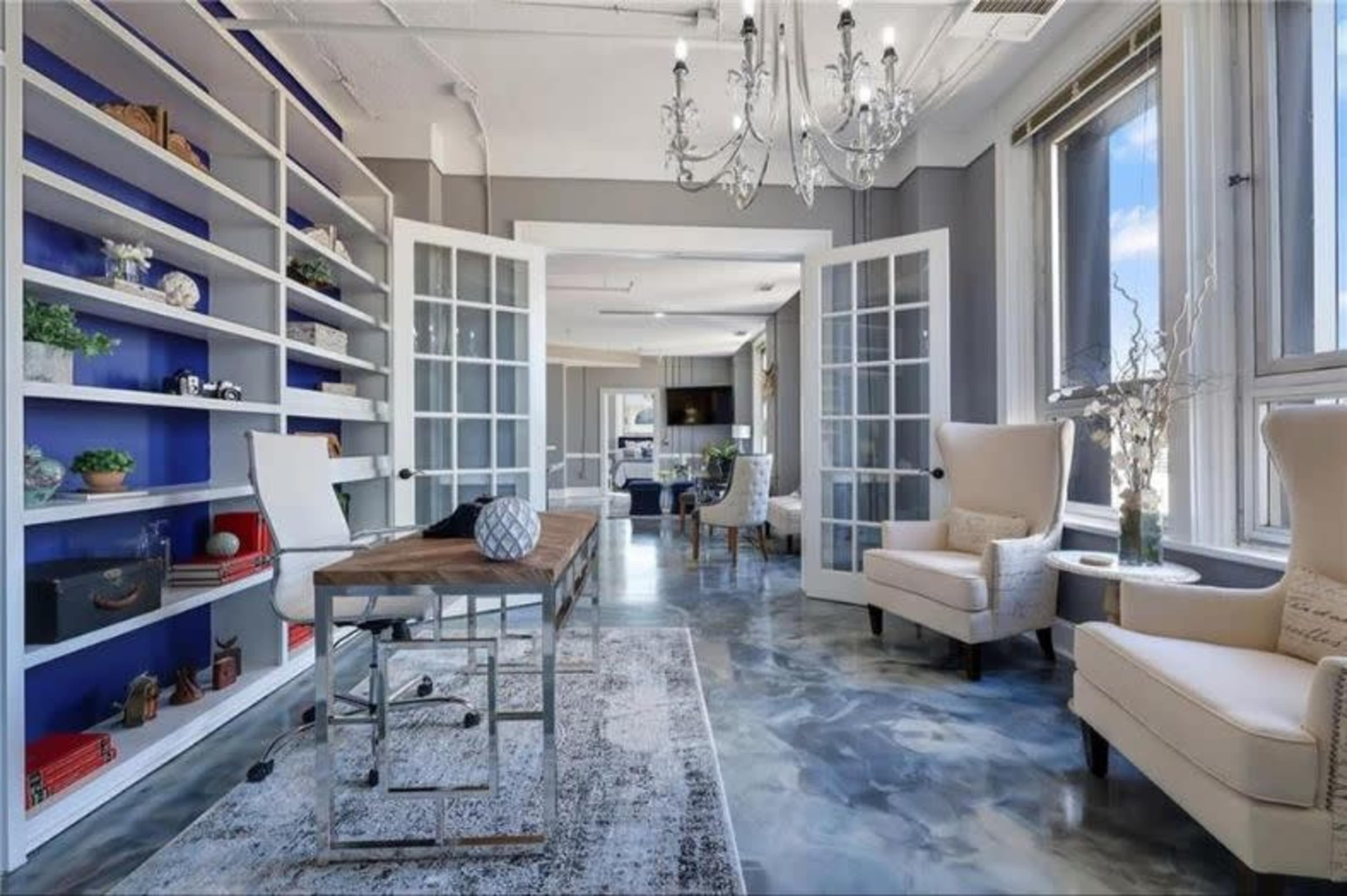 A bright room features a large bookshelf, a chandelier, and comfortable seating with a view through large windows.