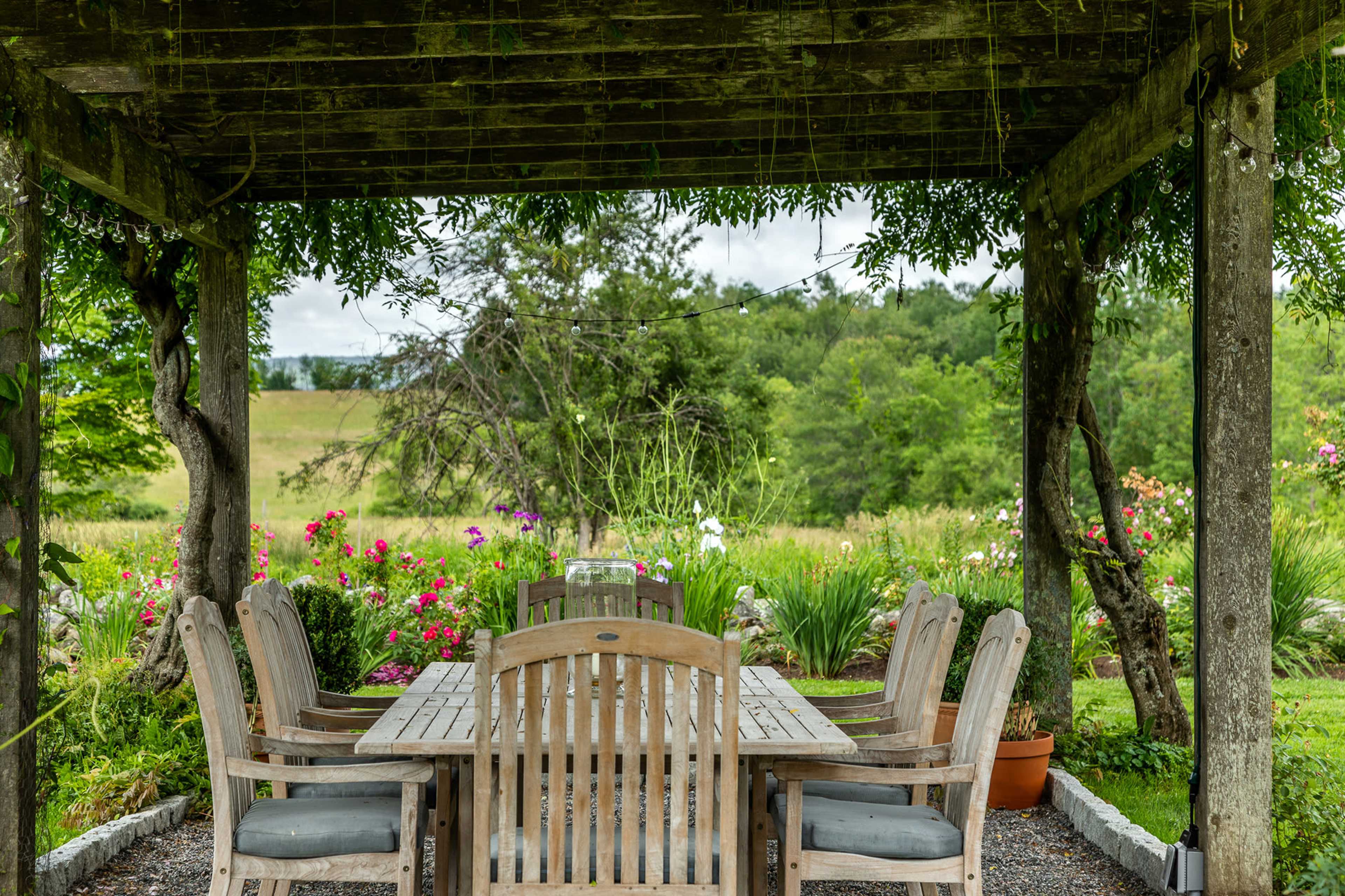 A wooden dining table surrounded by chairs is set under a rustic pergola, with a backdrop of lush greenery and blooming flowers.