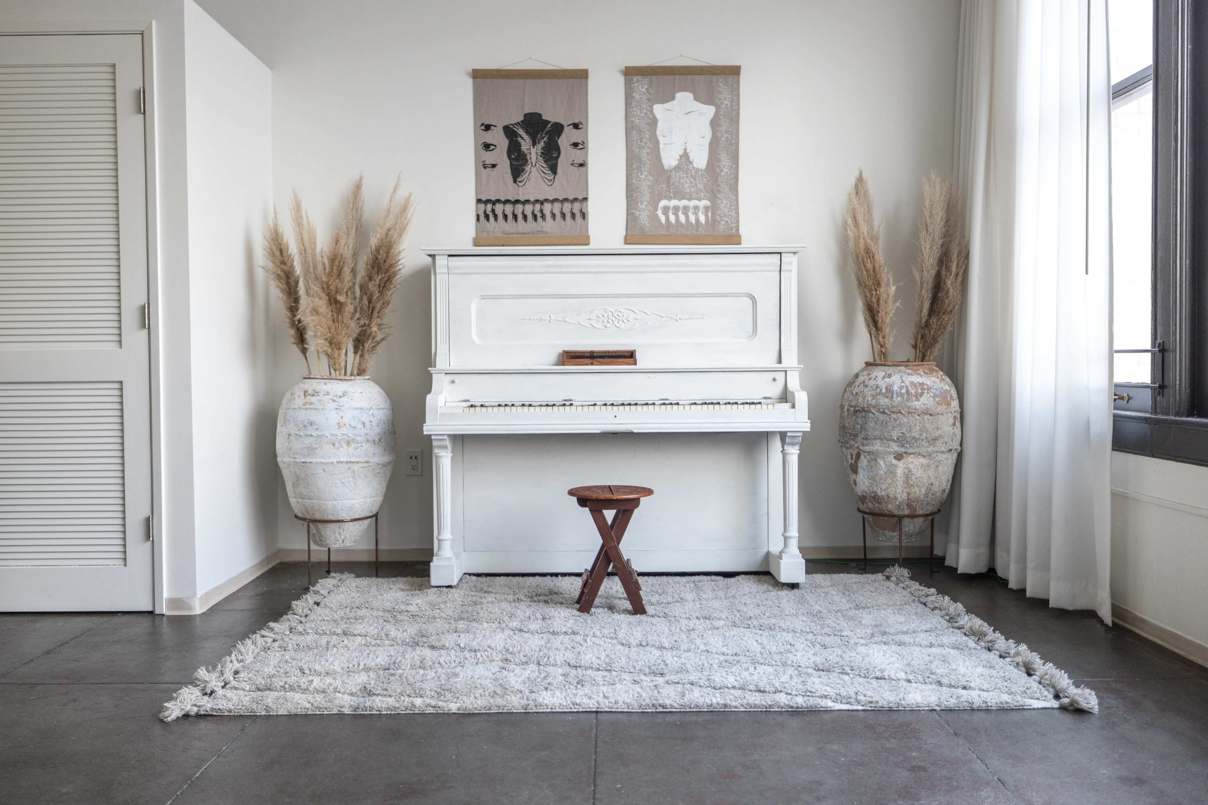 A white piano is positioned against a wall, accompanied by two large planters and a small wooden stool on a textured rug.