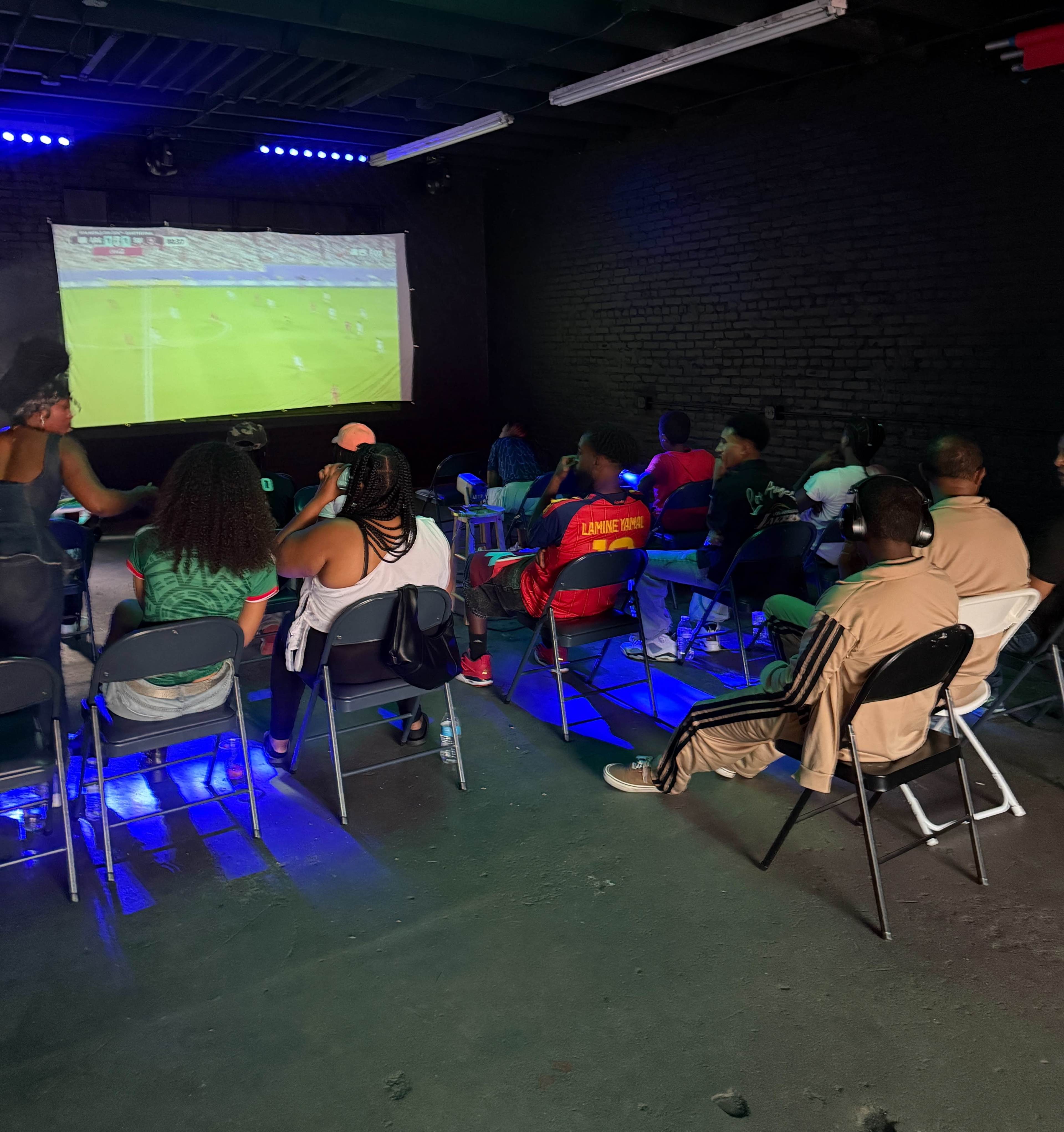 A group of people is seated in chairs, attentively watching a soccer match projected on a screen in a dimly lit room.