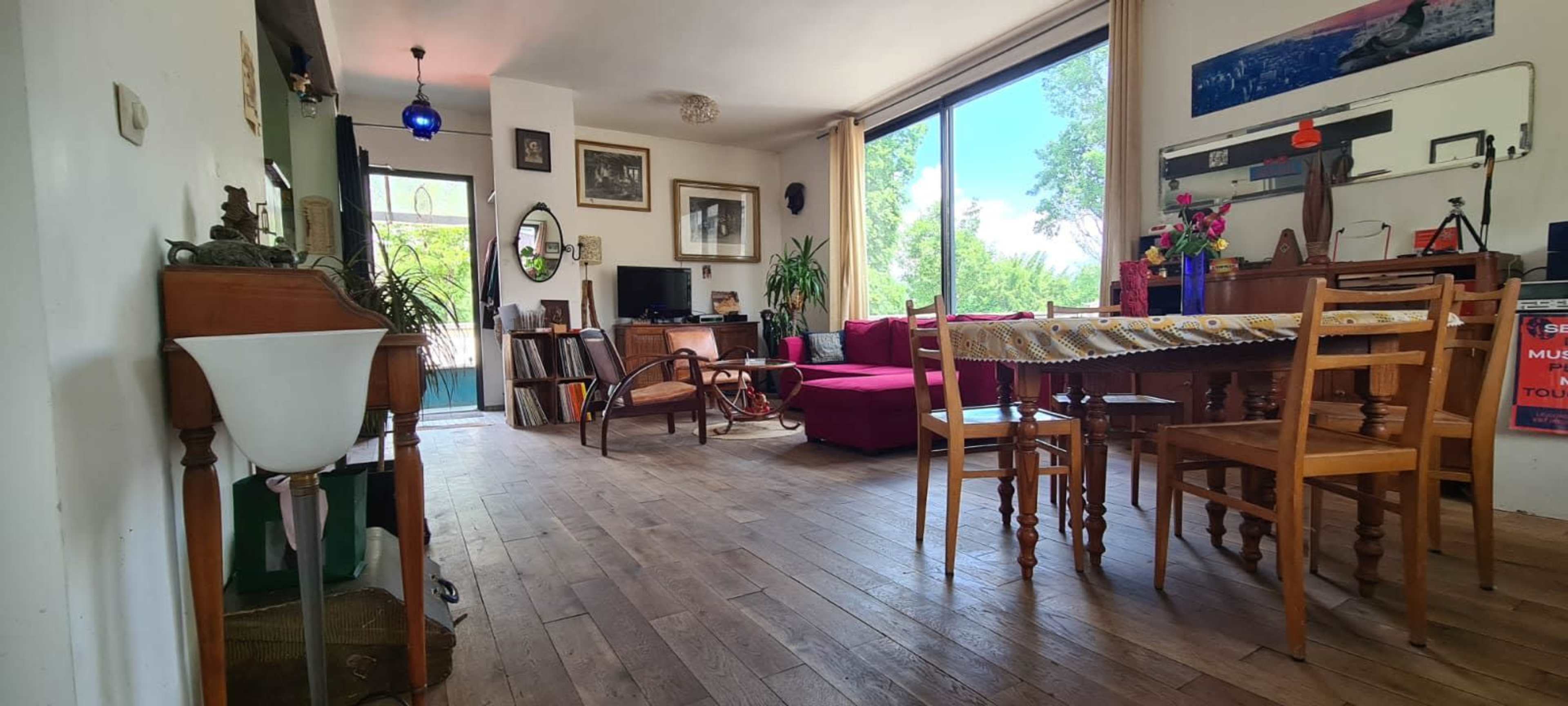 The image depicts a living space with a wooden floor, a dining table with chairs, a sofa, plants, and an array of decor against the walls.
