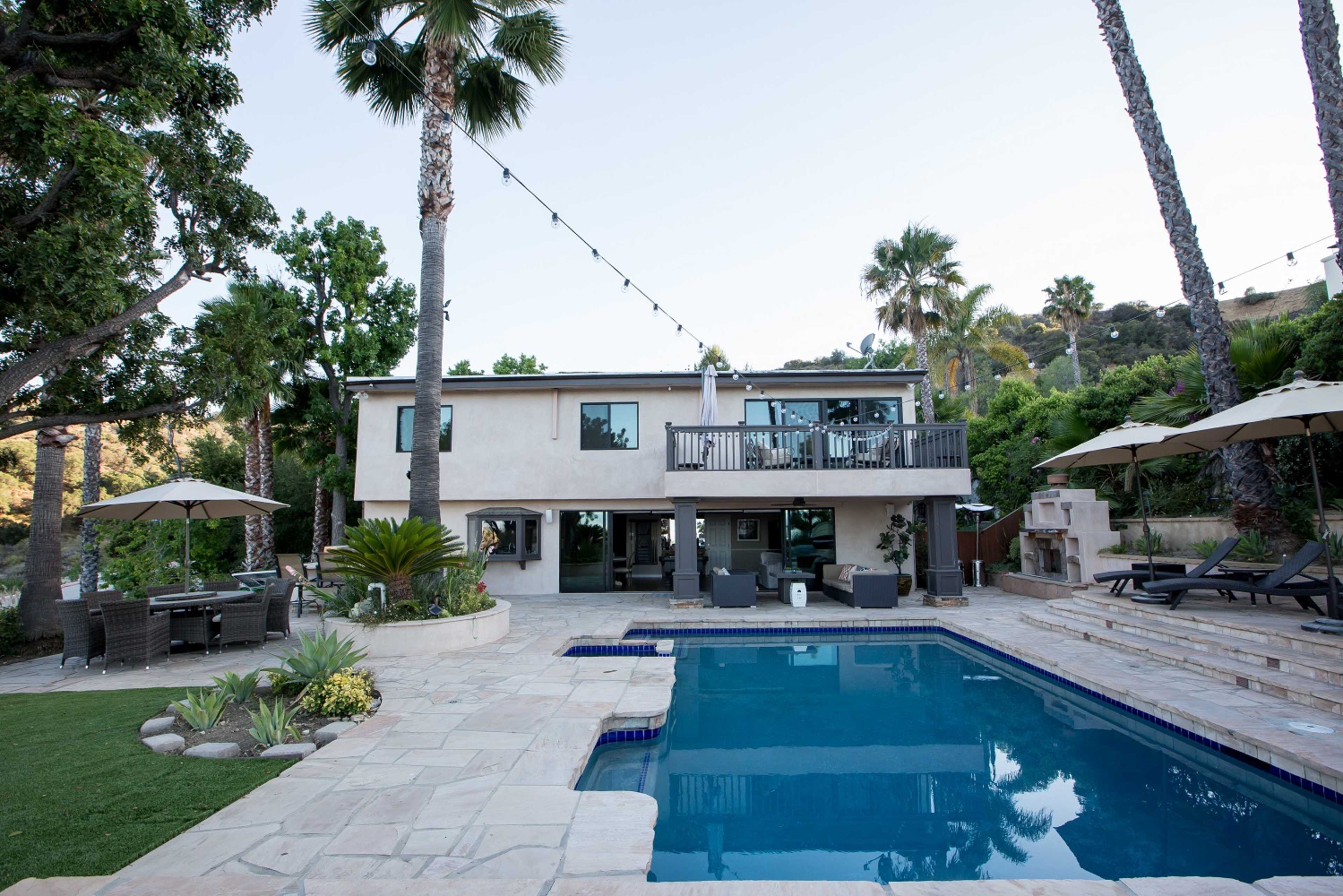 A two-story house with a swimming pool and outdoor seating area is surrounded by palm trees and landscaped greenery.