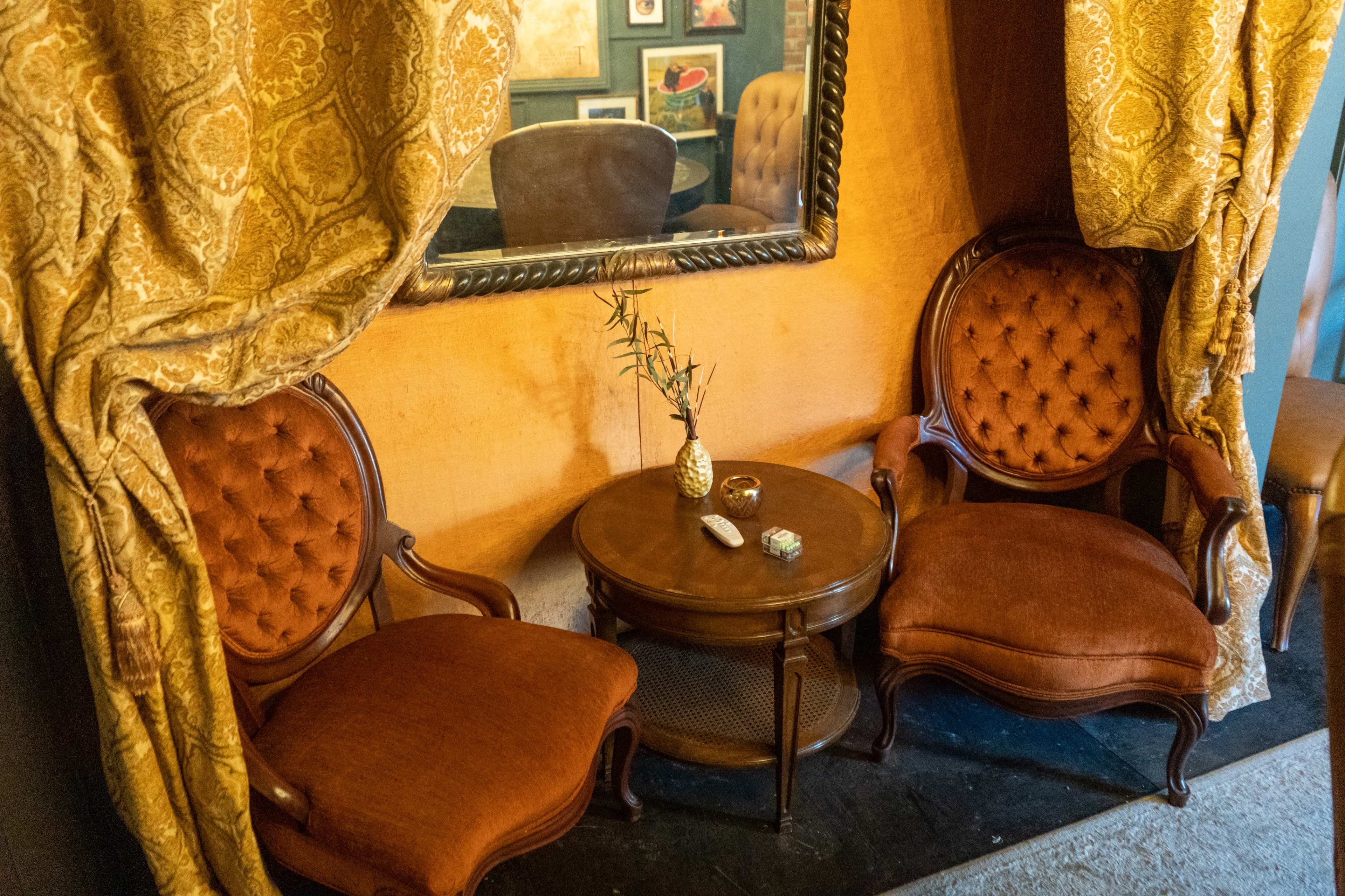 The image shows a cozy sitting area with two upholstered chairs and a small circular table, framed by decorative curtains and a mirror.