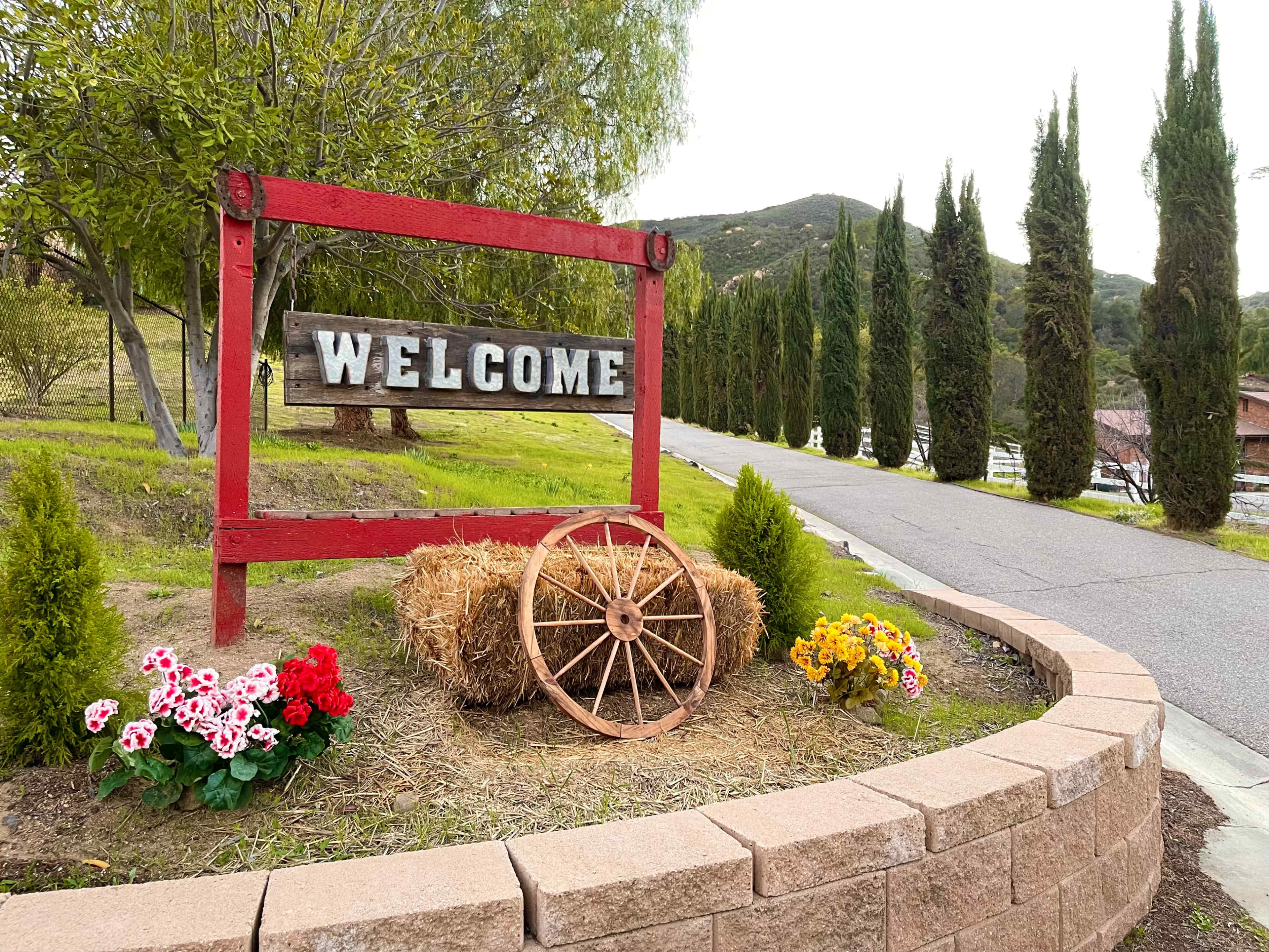 A red wooden welcome sign stands at the entrance of a landscaped area, surrounded by flowers and a wagon wheel along a paved road lined with tall cypress trees.