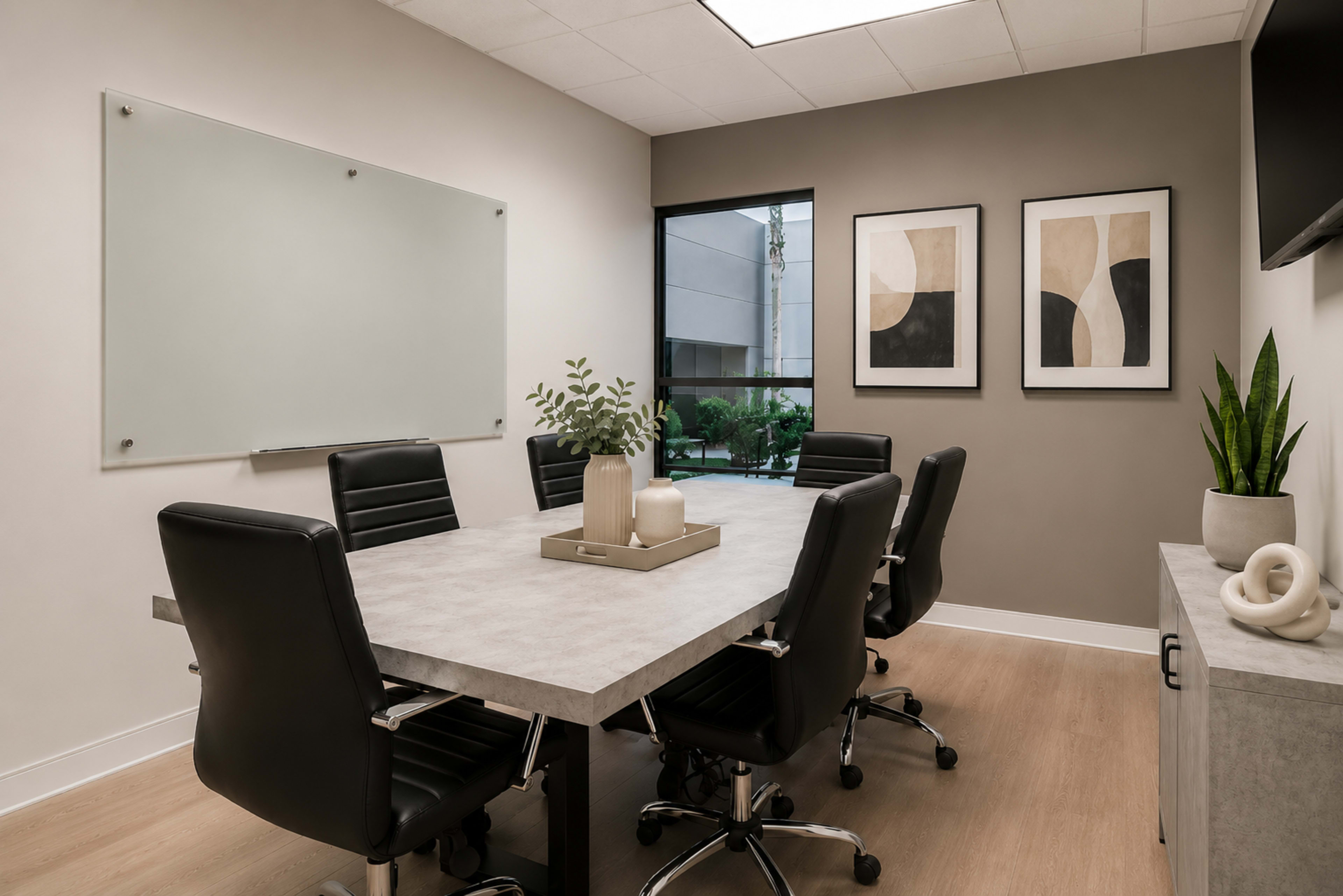 A modern conference room features a large table surrounded by black leather chairs, with framed artwork on the walls and a whiteboard on one side.