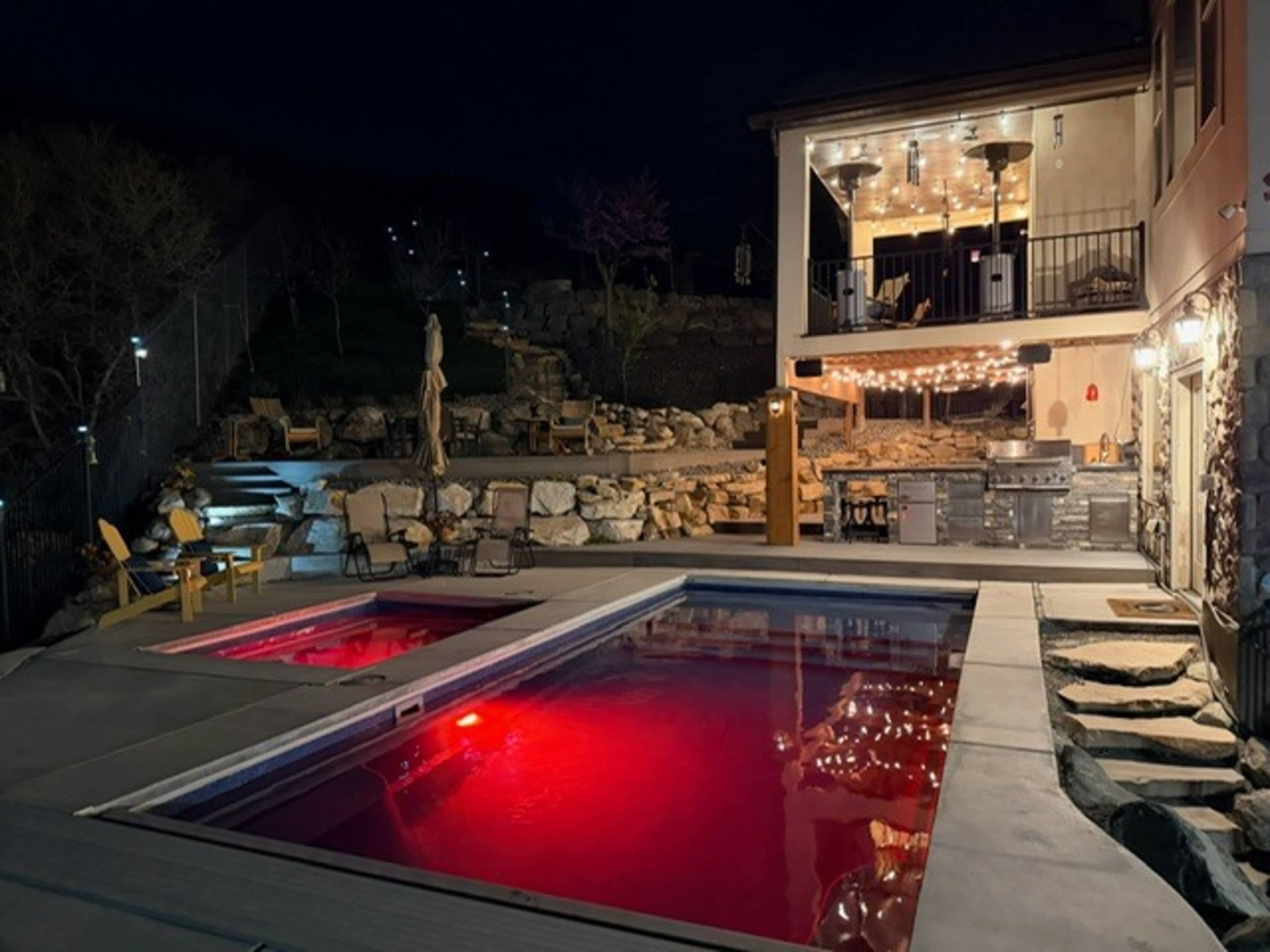 A lit backyard features a pool with red lighting, surrounded by stone landscaping and a house with a balcony above.