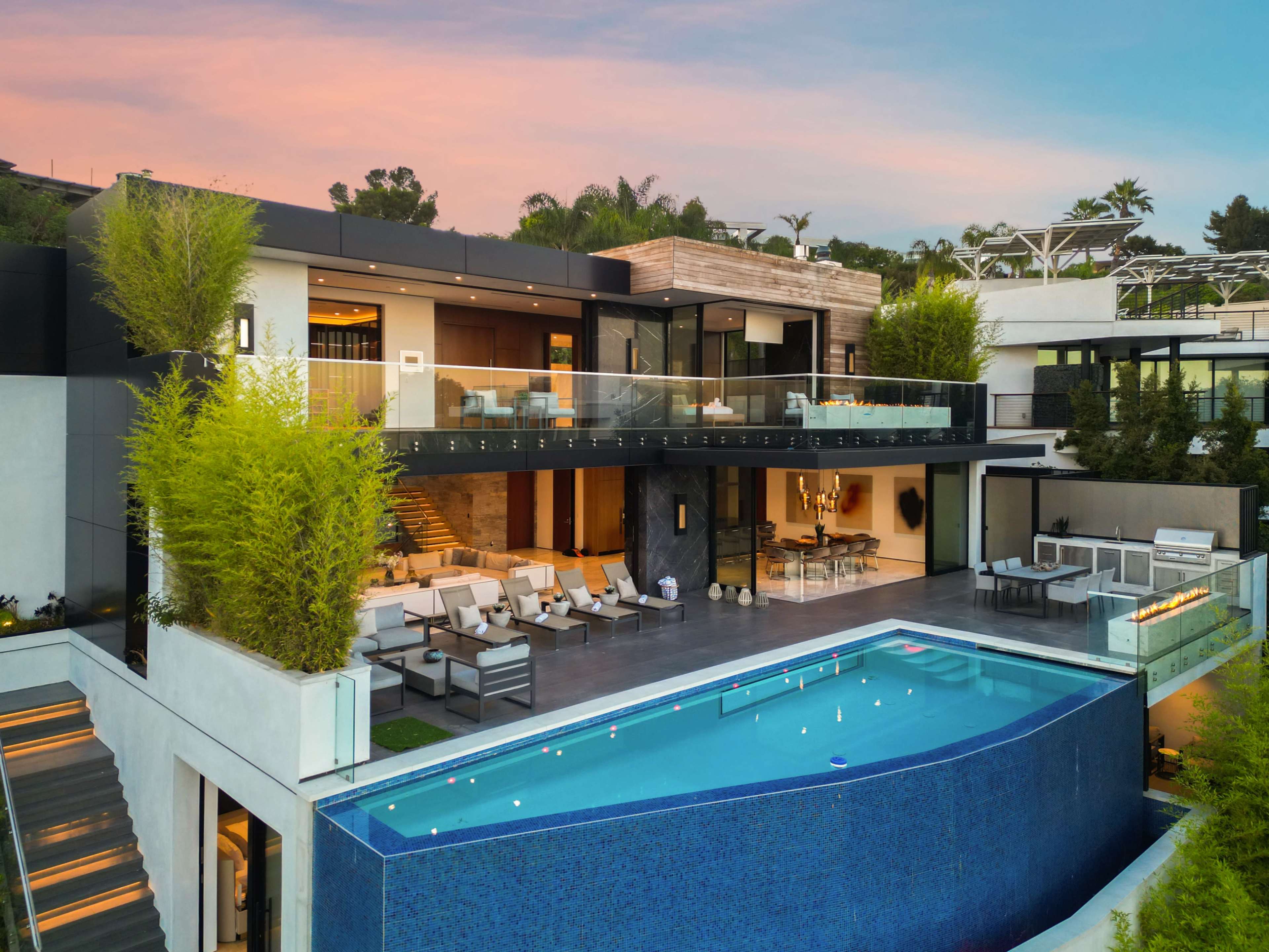 The image shows a modern villa with a sleek design, featuring a large swimming pool and multiple lounge areas surrounded by greenery.