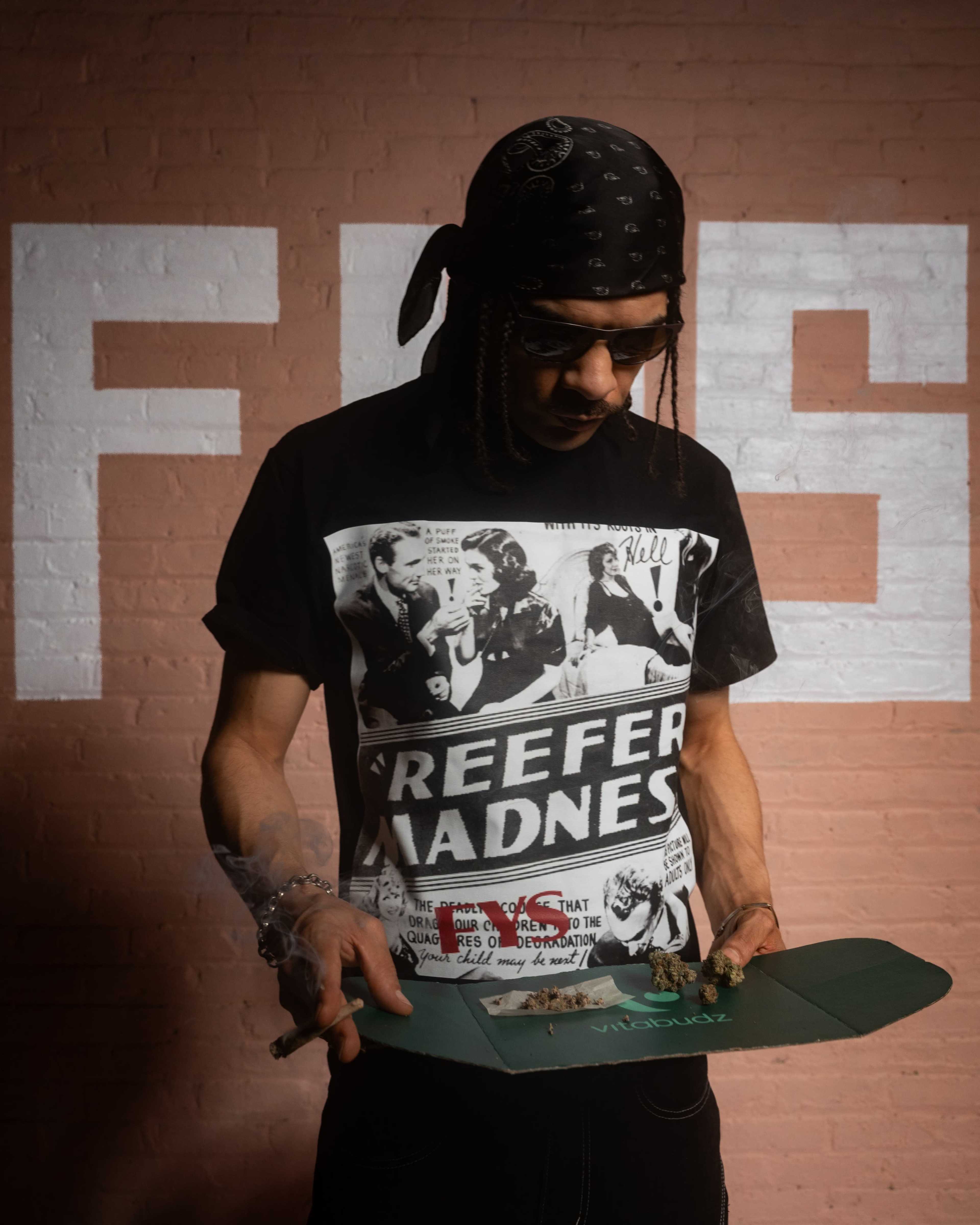 A person wearing a "Reefer Madness" T-shirt stands holding a rolling tray and a blunt in a room with a pink brick wall featuring large text.