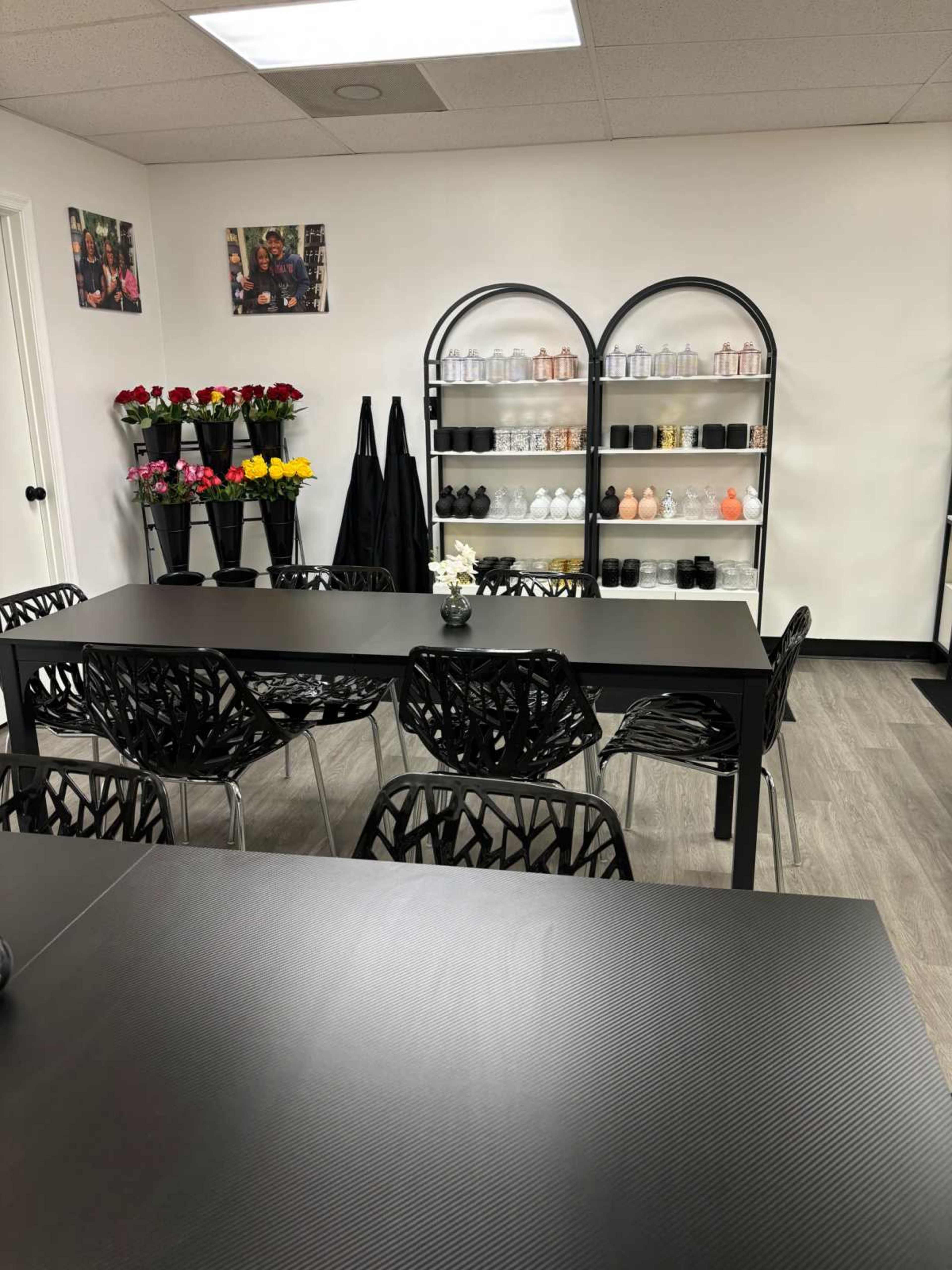 A modern classroom or meeting space with black tables and chairs, a display shelf with various bottles, and floral arrangements on the wall.