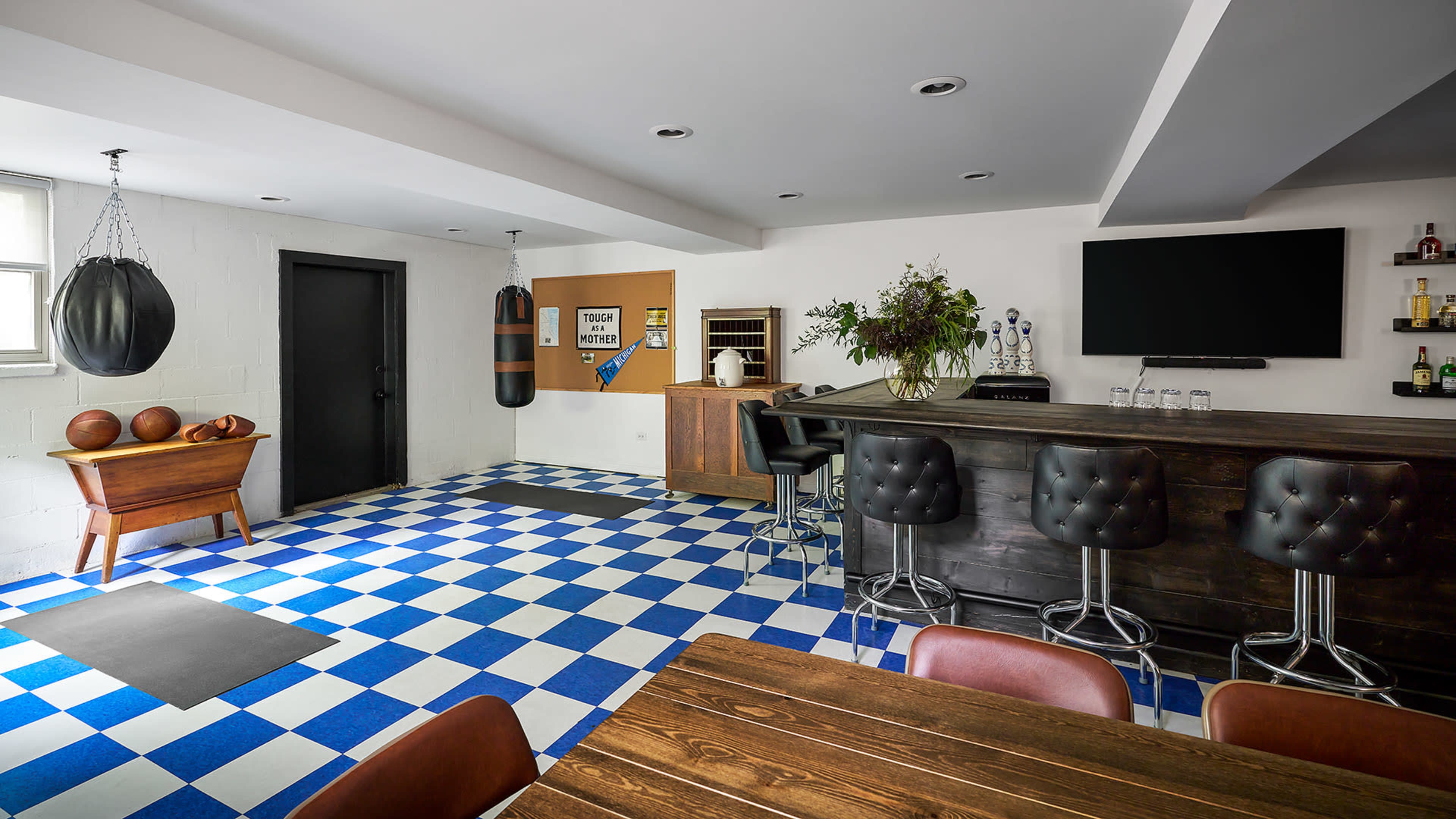 A basement room featuring a boxing bag, a bar area, a dining table, and a checkered blue and white tiled floor.
