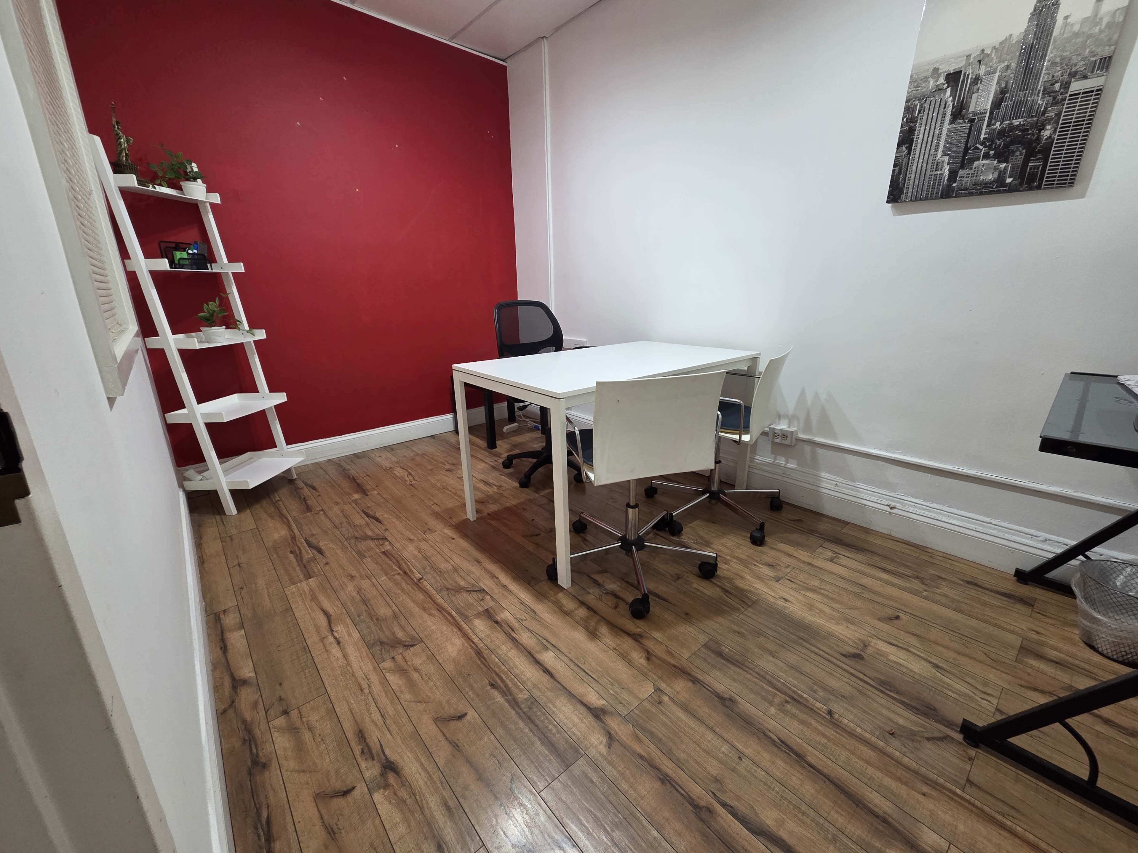 The image shows a small office space with a red accent wall, a white table surrounded by chairs, and a shelf in the corner.