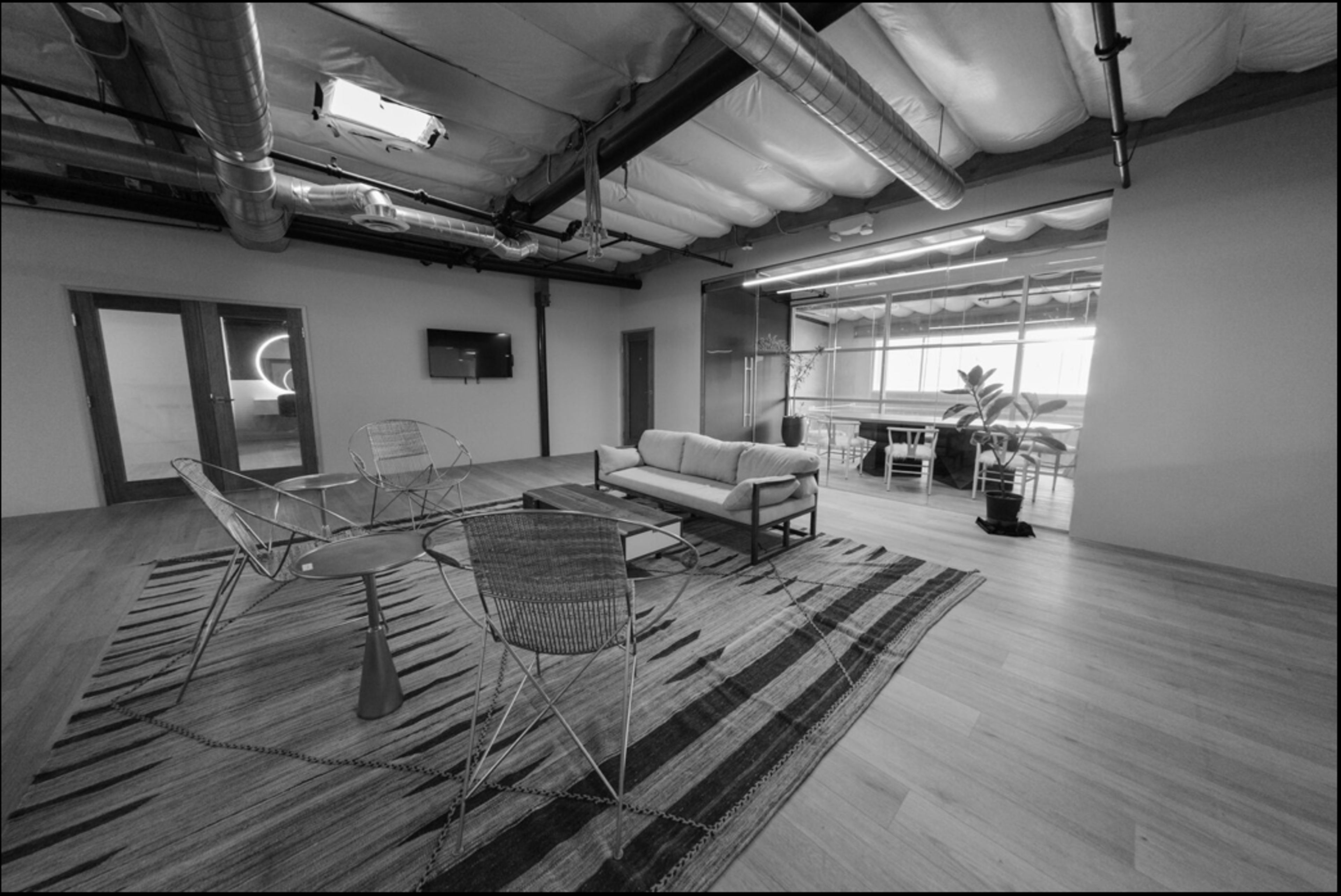 A modern office space featuring a seating area with three wire chairs, a sofa, a round table on a patterned rug, and large windows in the background.