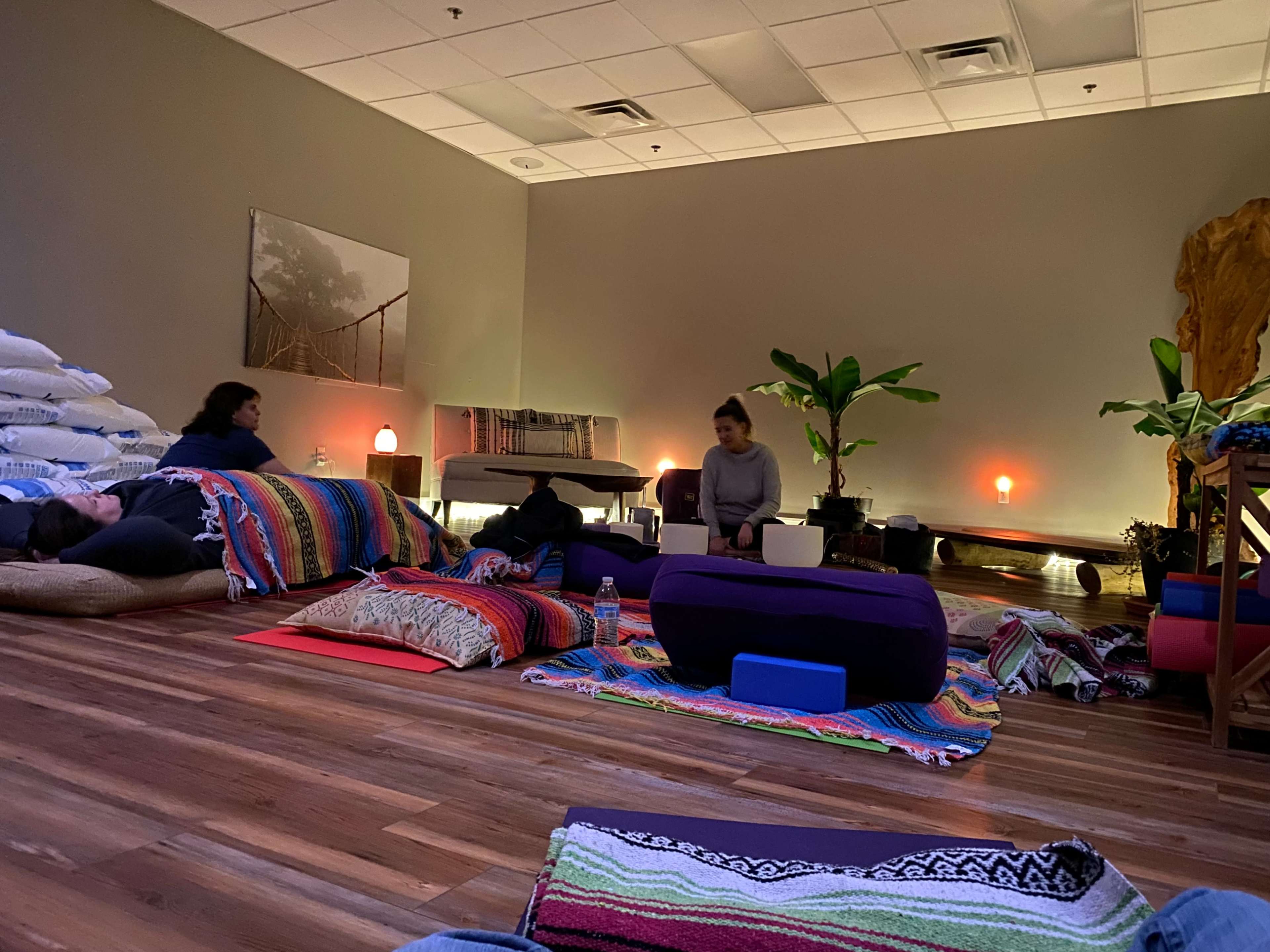 A dimly lit room with people sitting and lying on colorful blankets and cushions, surrounded by plants and soft lighting.