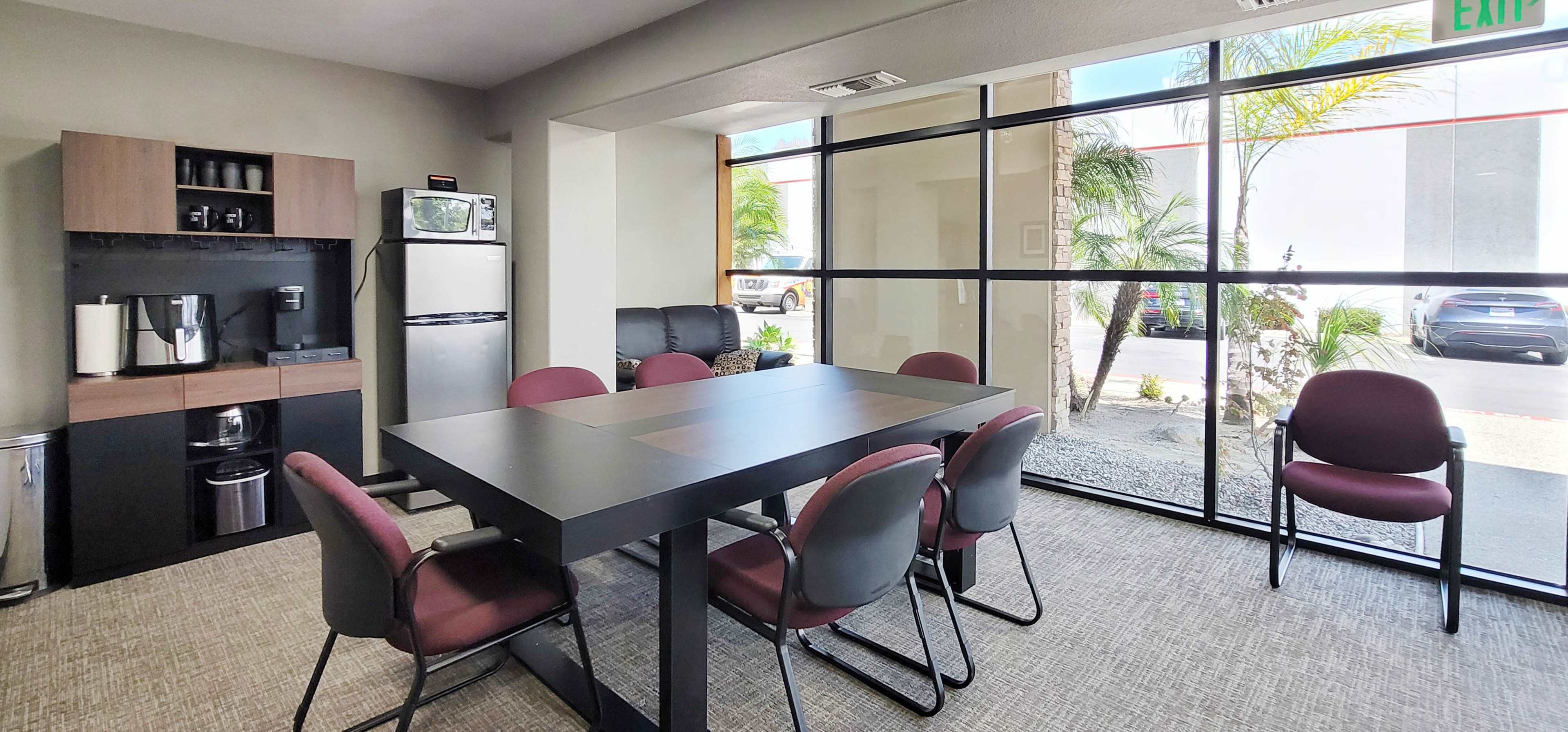 The image shows a small meeting room with a black table surrounded by six maroon chairs, a kitchenette area, and large windows allowing natural light.