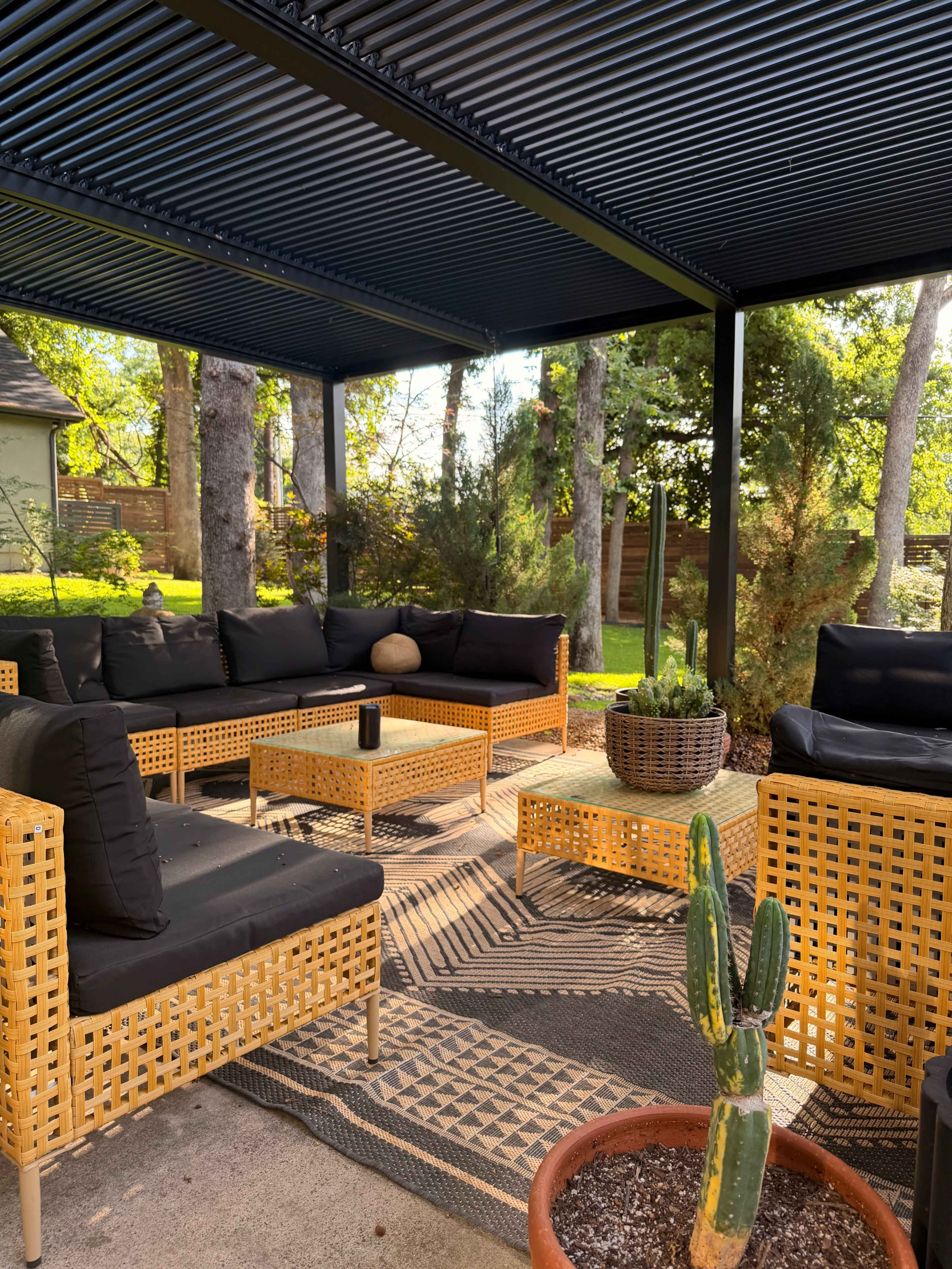 The image shows a shaded outdoor living area with wicker furniture arranged around a coffee table, surrounded by trees and potted plants.