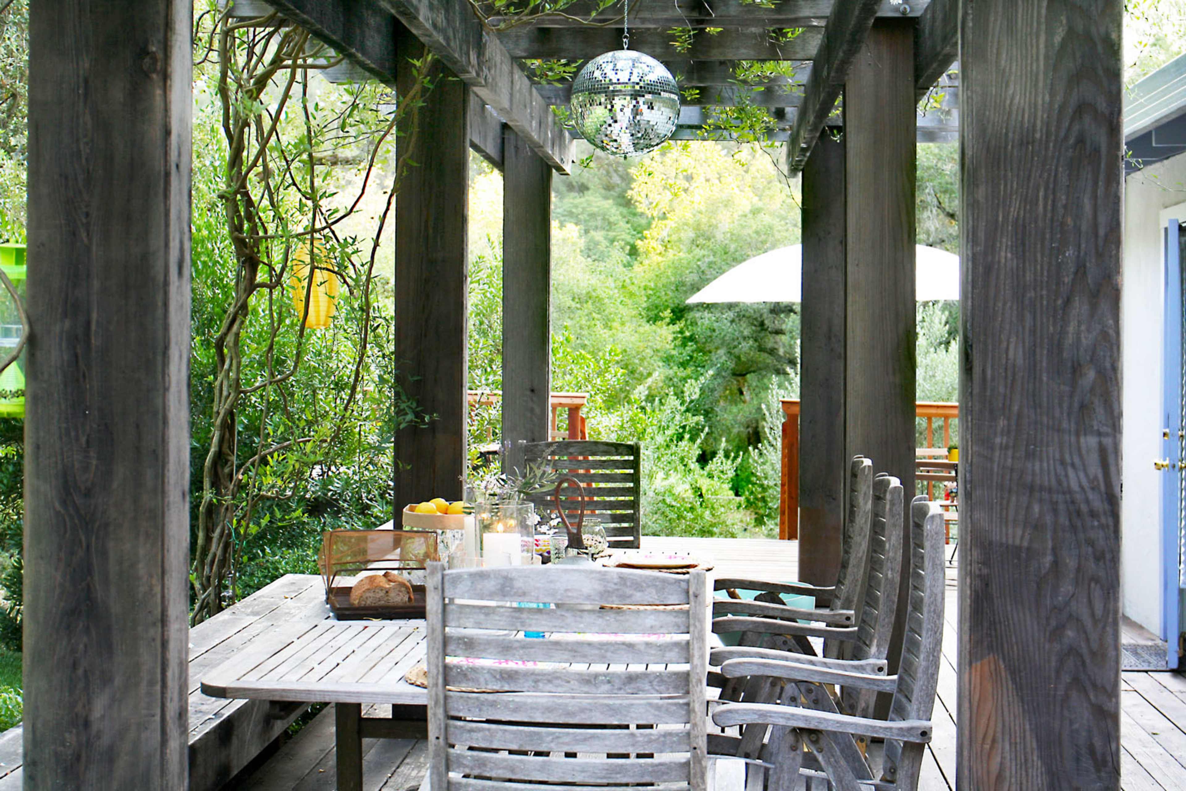 A wooden table with several chairs is set under a pergola surrounded by greenery and illuminated by a hanging disco ball.