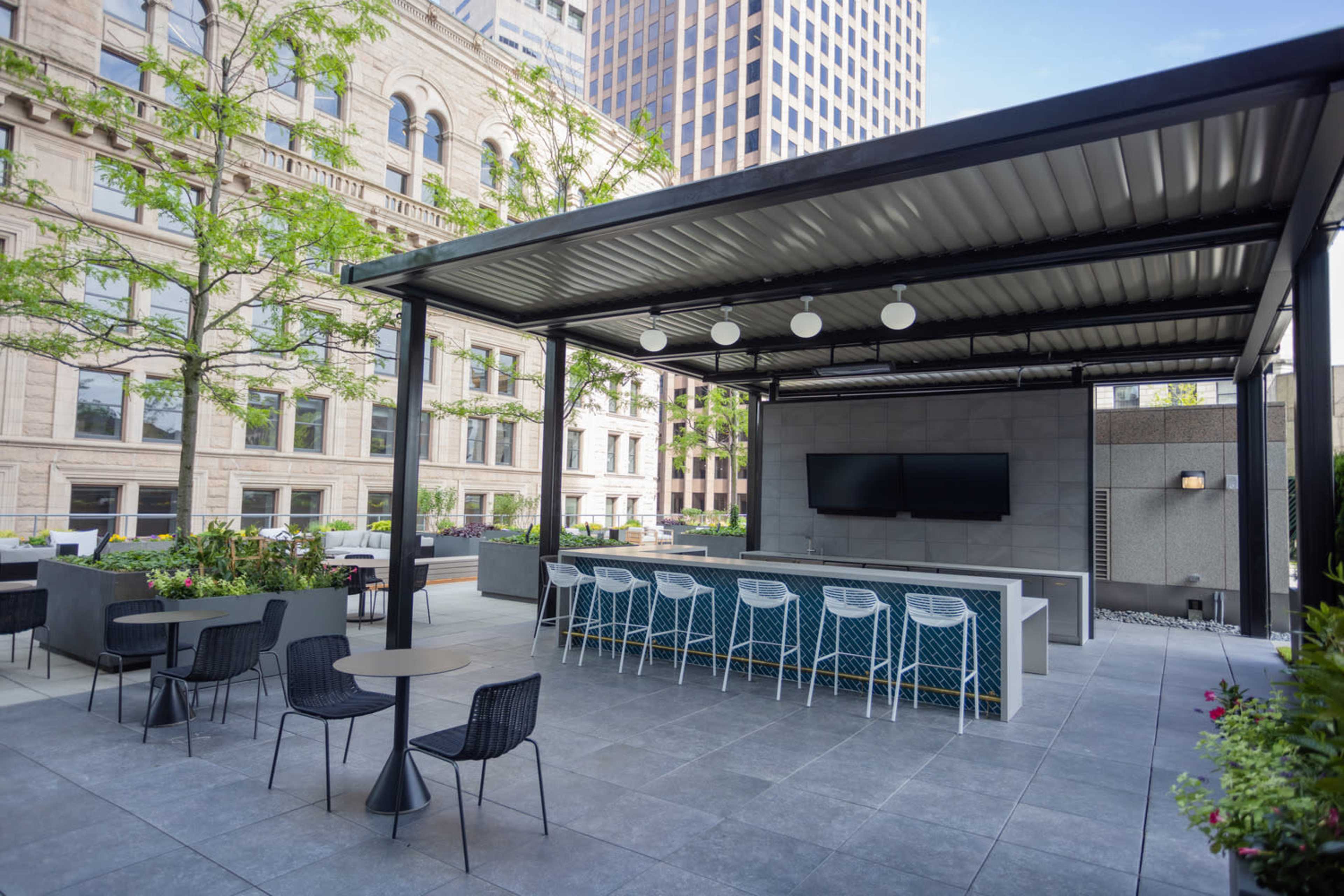 An outdoor patio area with a covered bar, seating, and two television screens, surrounded by modern landscaping and buildings.