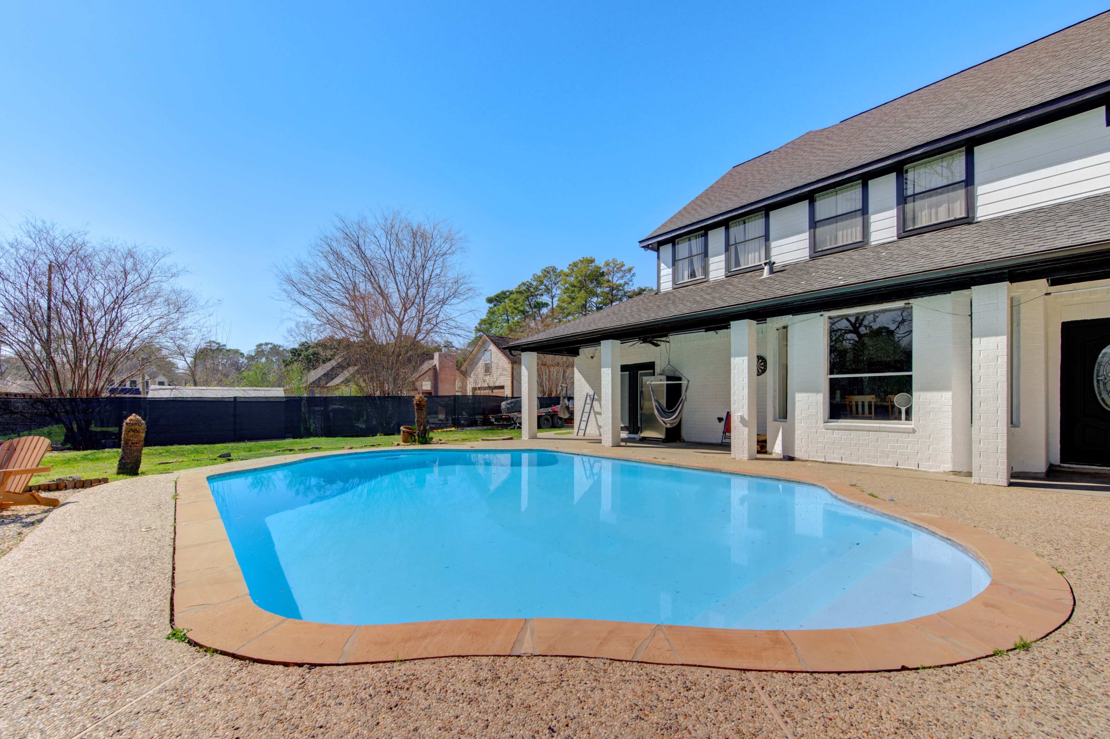 A backyard features a rectangular swimming pool surrounded by a concrete patio and a two-story house with a black roof and white walls.