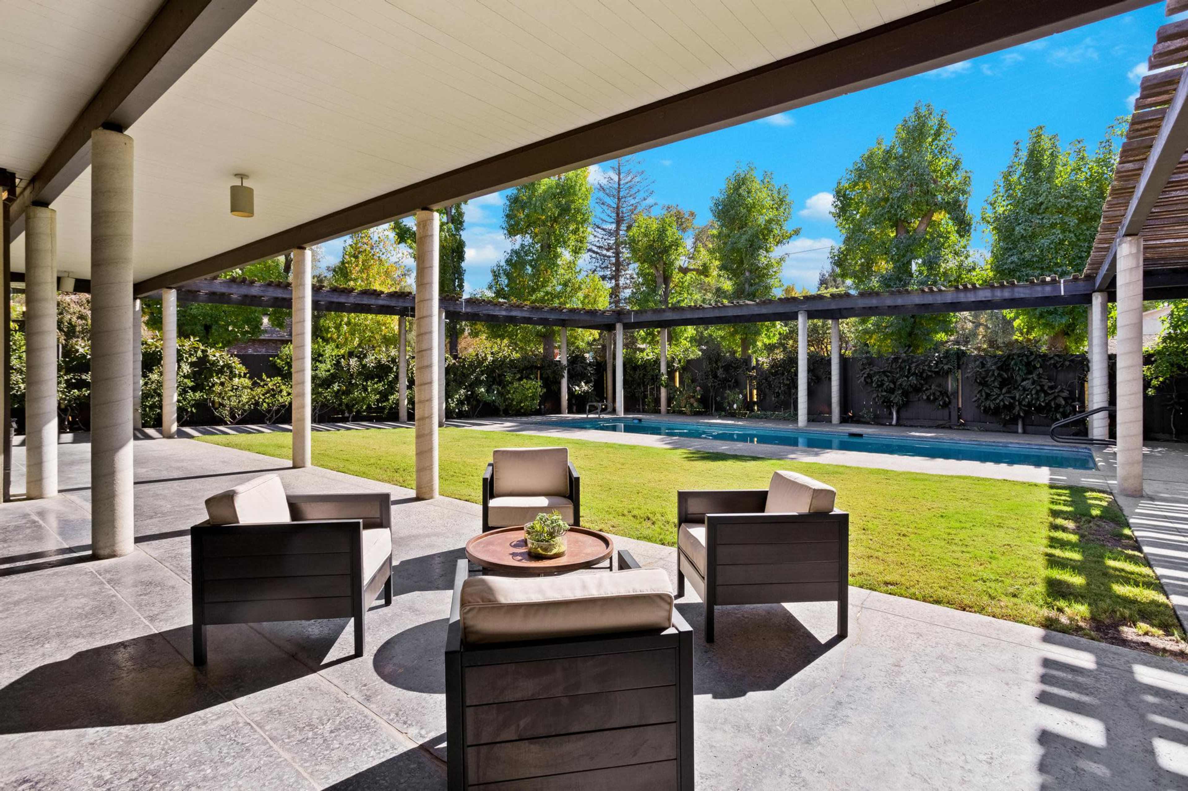 A patio area with four chairs arranged around a small table, overlooking a swimming pool surrounded by greenery.