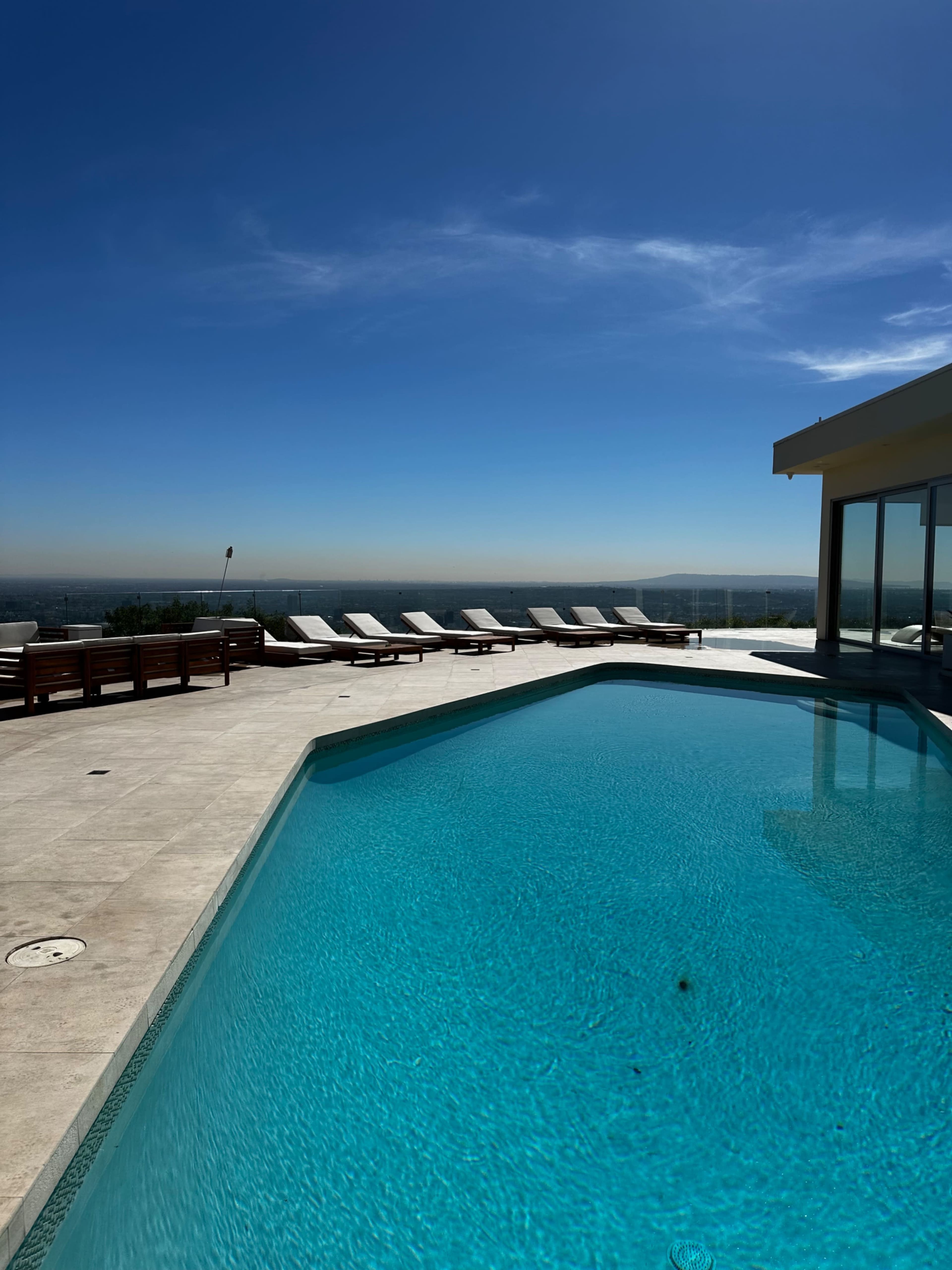 A clear blue swimming pool surrounded by lounge chairs and a panoramic view of the distant landscape under a bright sky.